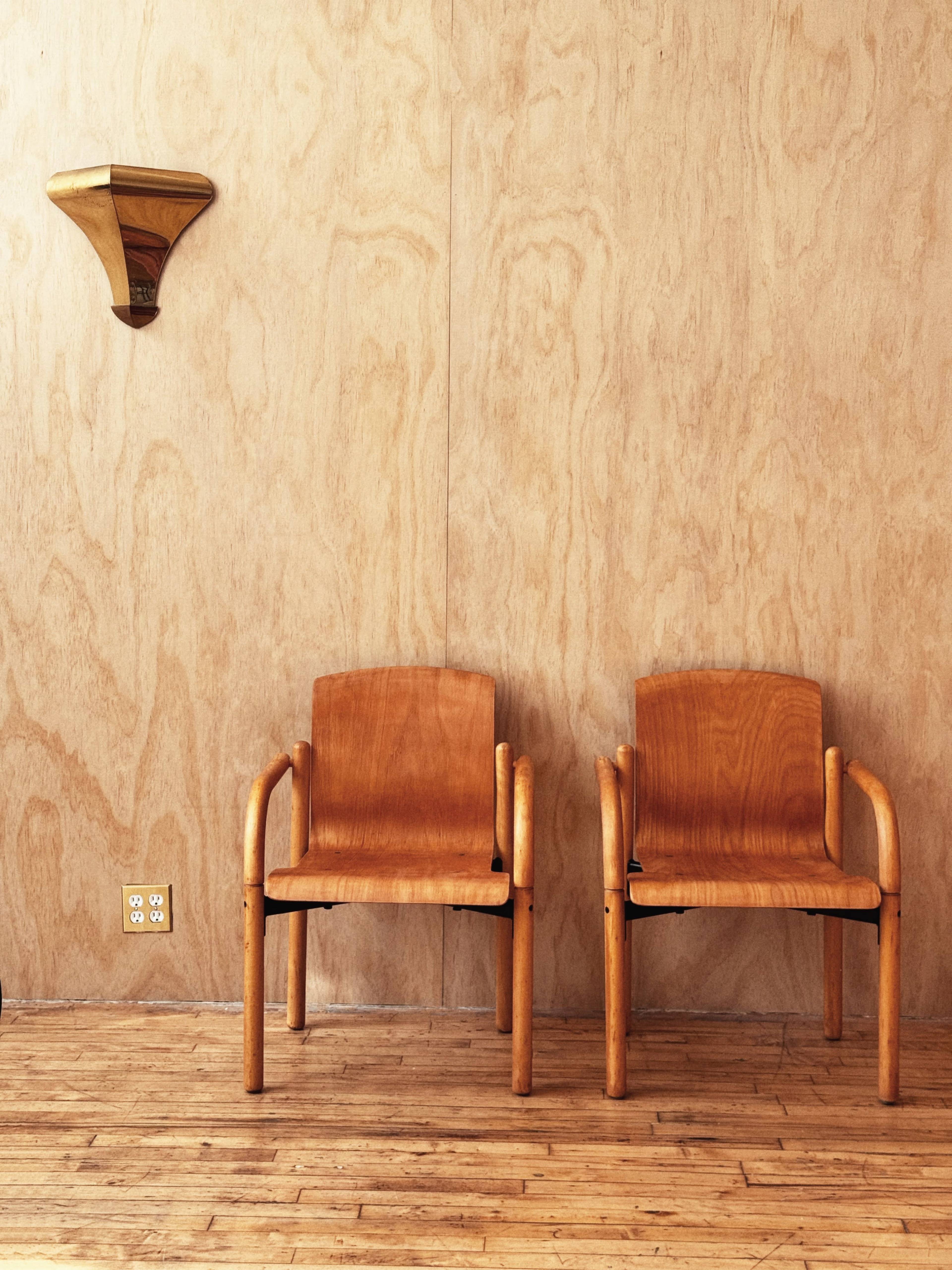 Two wooden chairs are positioned against a plywood wall, with a unique wall-mounted light fixture above them.