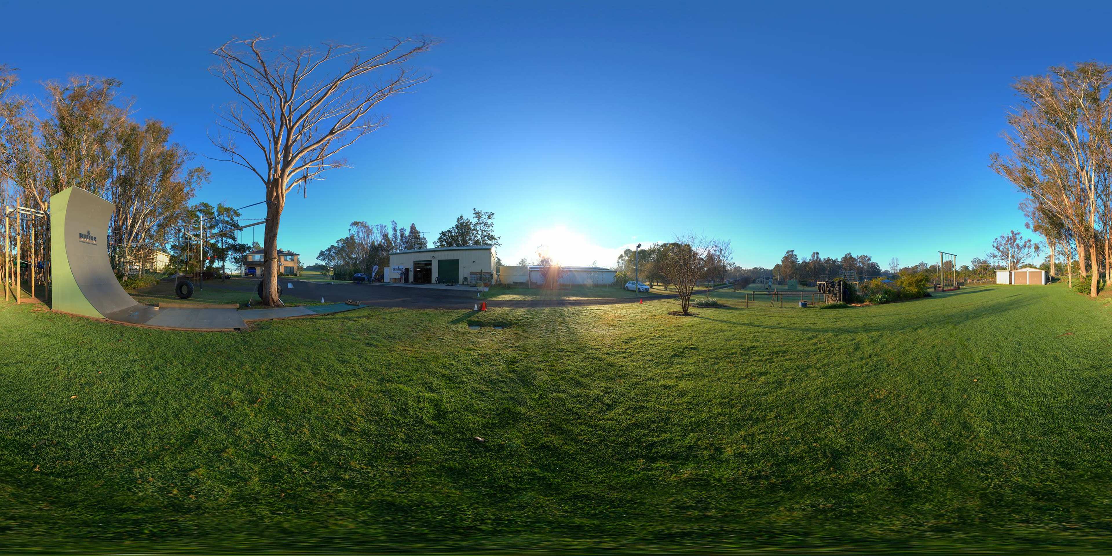 A panoramic view of a park-like area with a grass field, a half-pipe structure, and surrounded by trees and buildings under a clear blue sky.