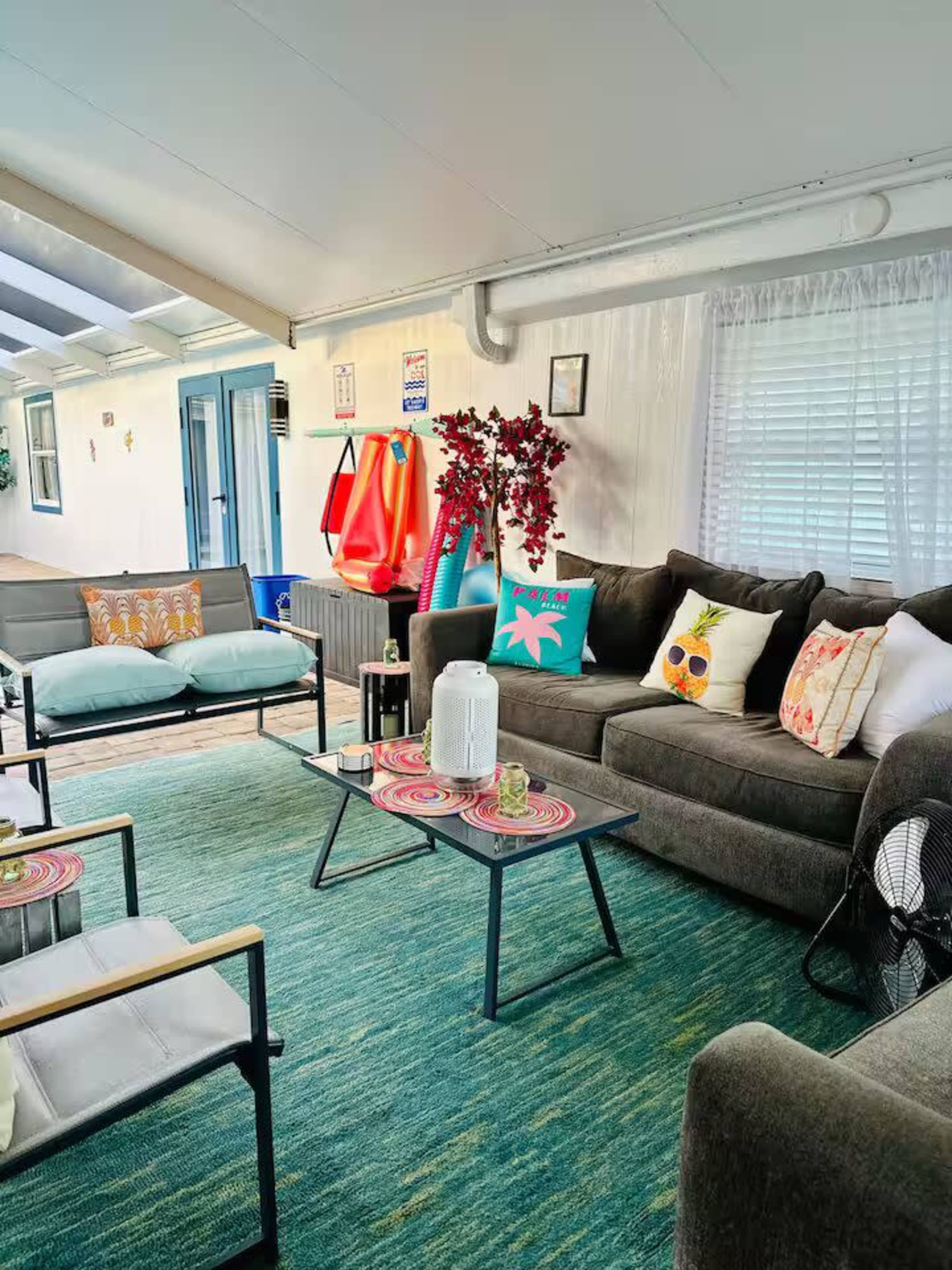 The image shows a cozy living area with a sofa, a small table, colorful cushions, a rug, and light streaming in through the windows.