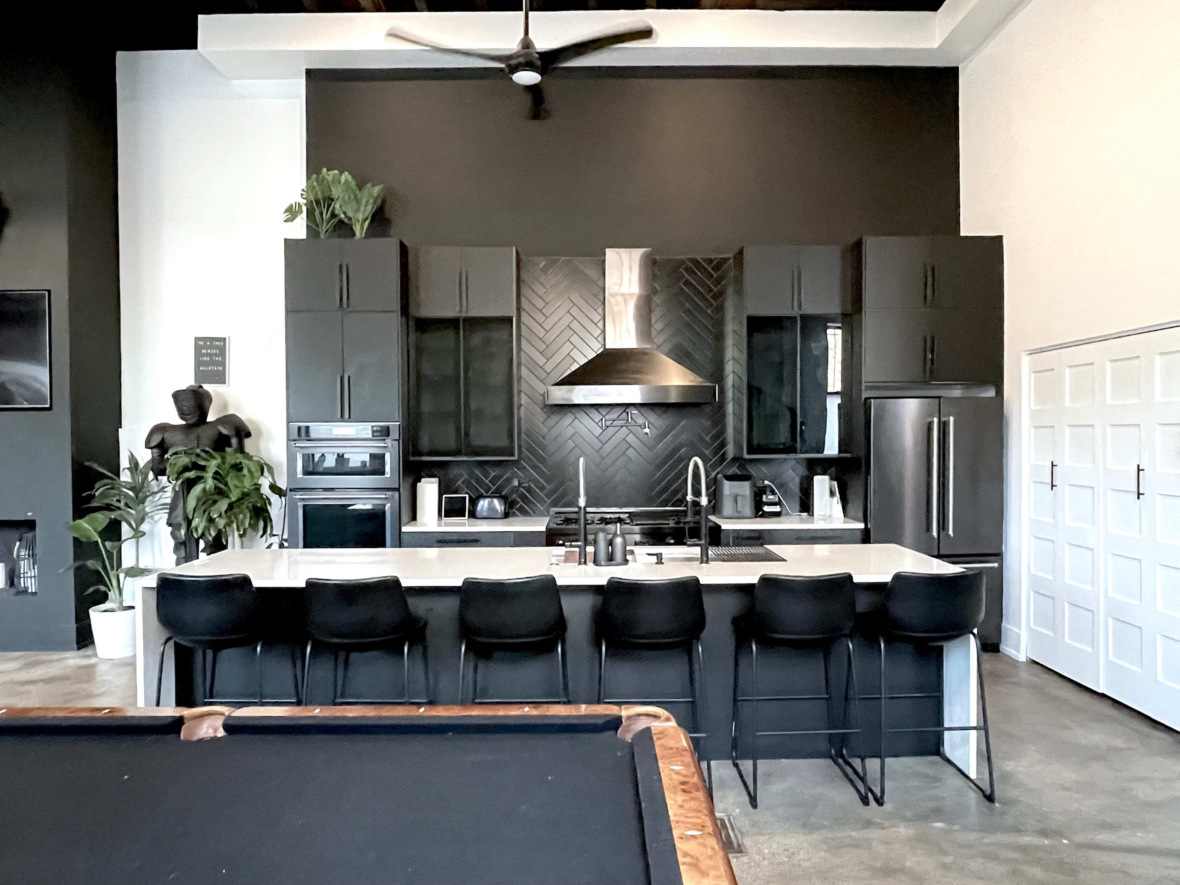 A modern kitchen with black cabinets, a large island with seating, and stainless steel appliances, complemented by a herringbone-patterned backsplash.