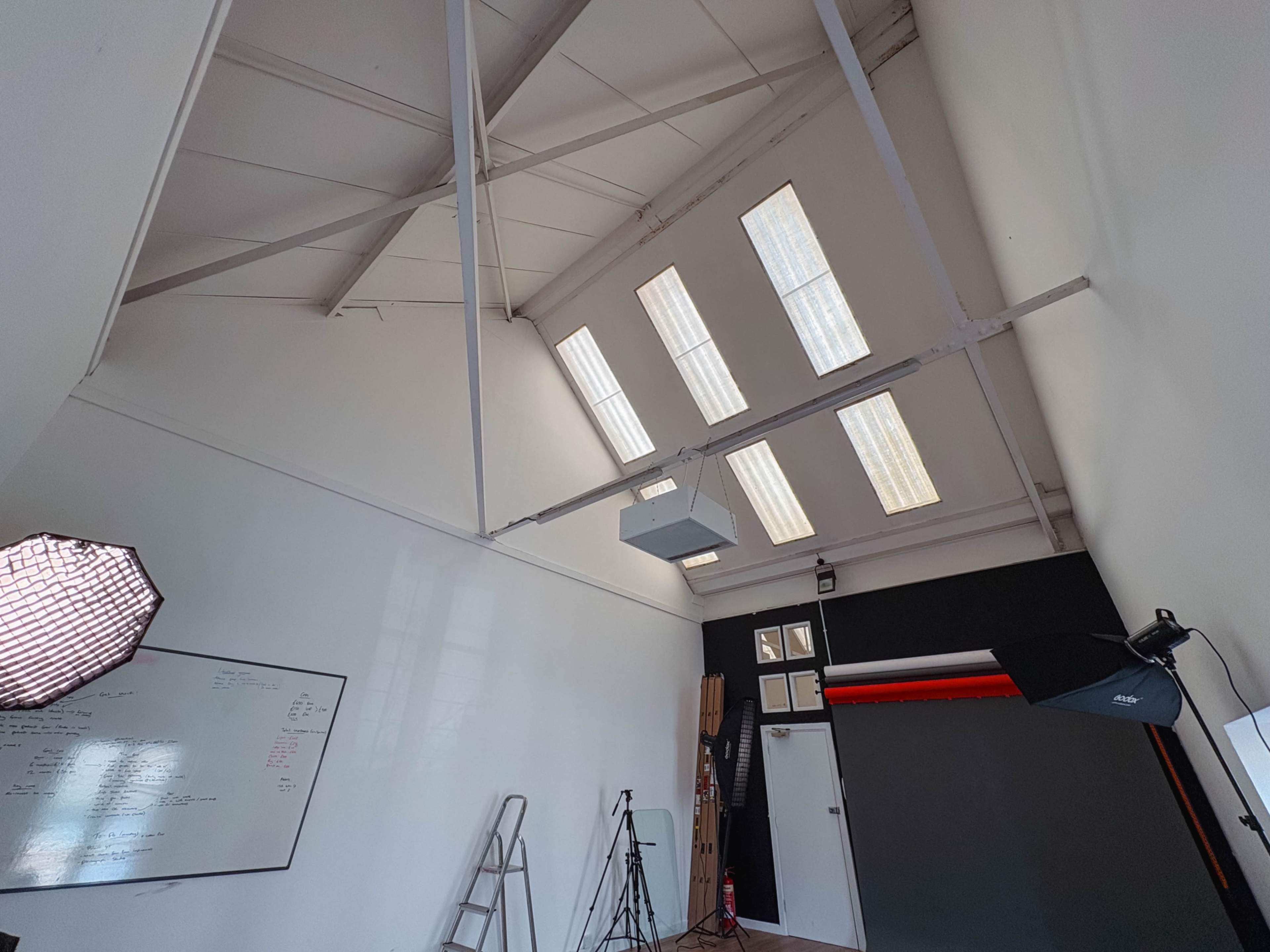 The image shows an interior space with high ceilings, large skylights, a whiteboard on one wall, and photography equipment set up to the side.