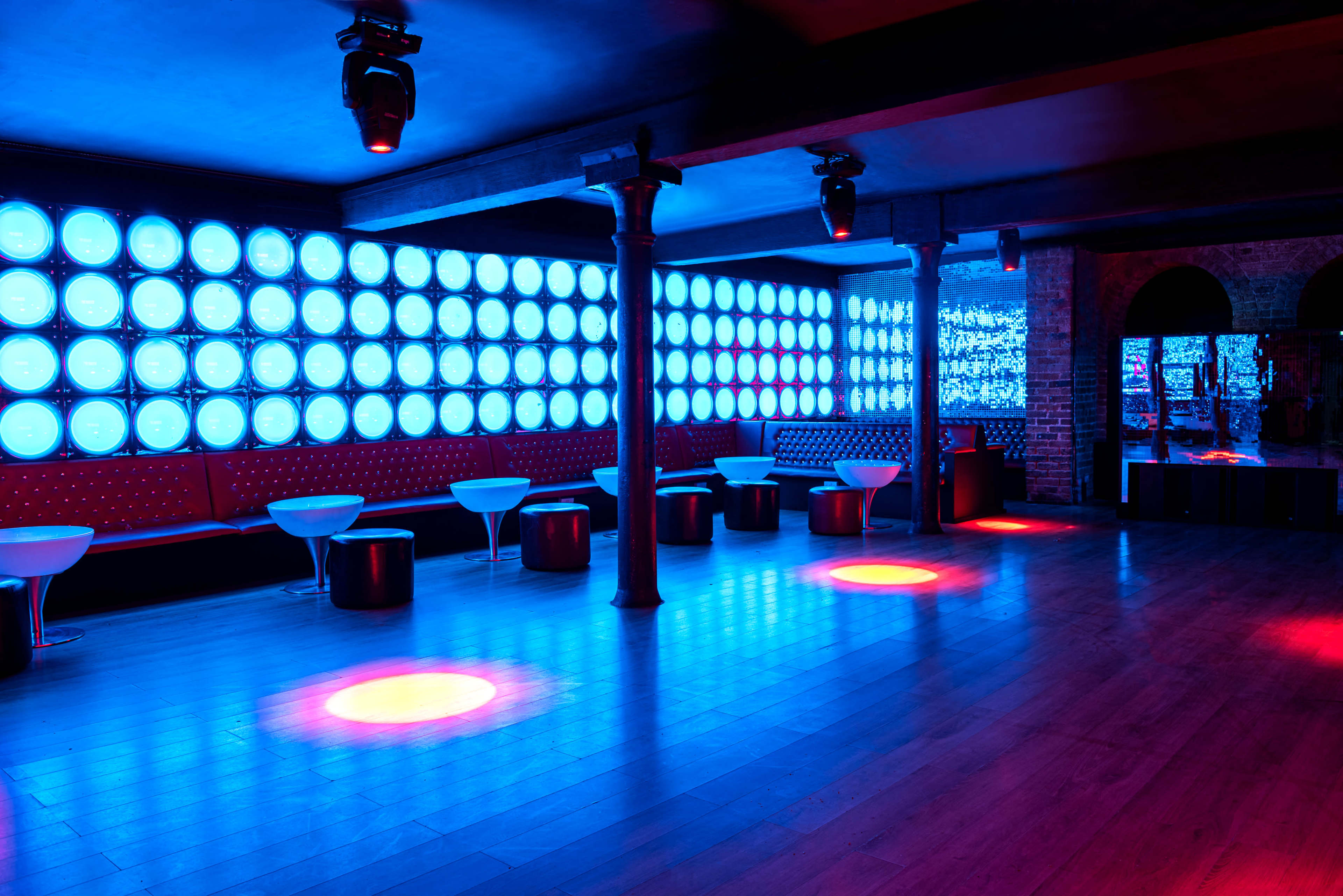 A dimly lit nightclub interior with blue and red lighting, featuring white lounge chairs and tables arranged around a wooden floor.