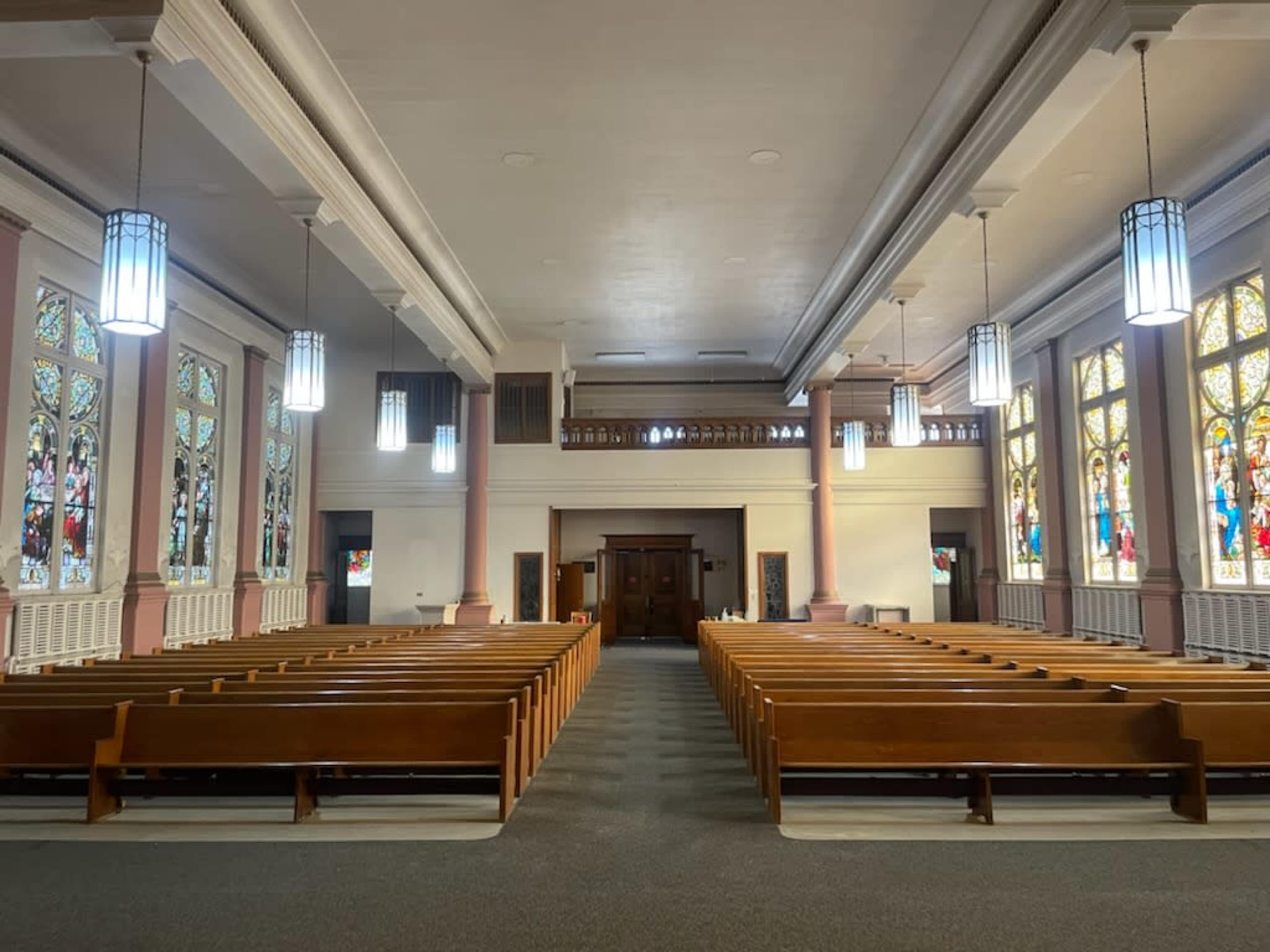 The interior of a church with rows of wooden pews facing a wooden altar, surrounded by stained glass windows and modern light fixtures.