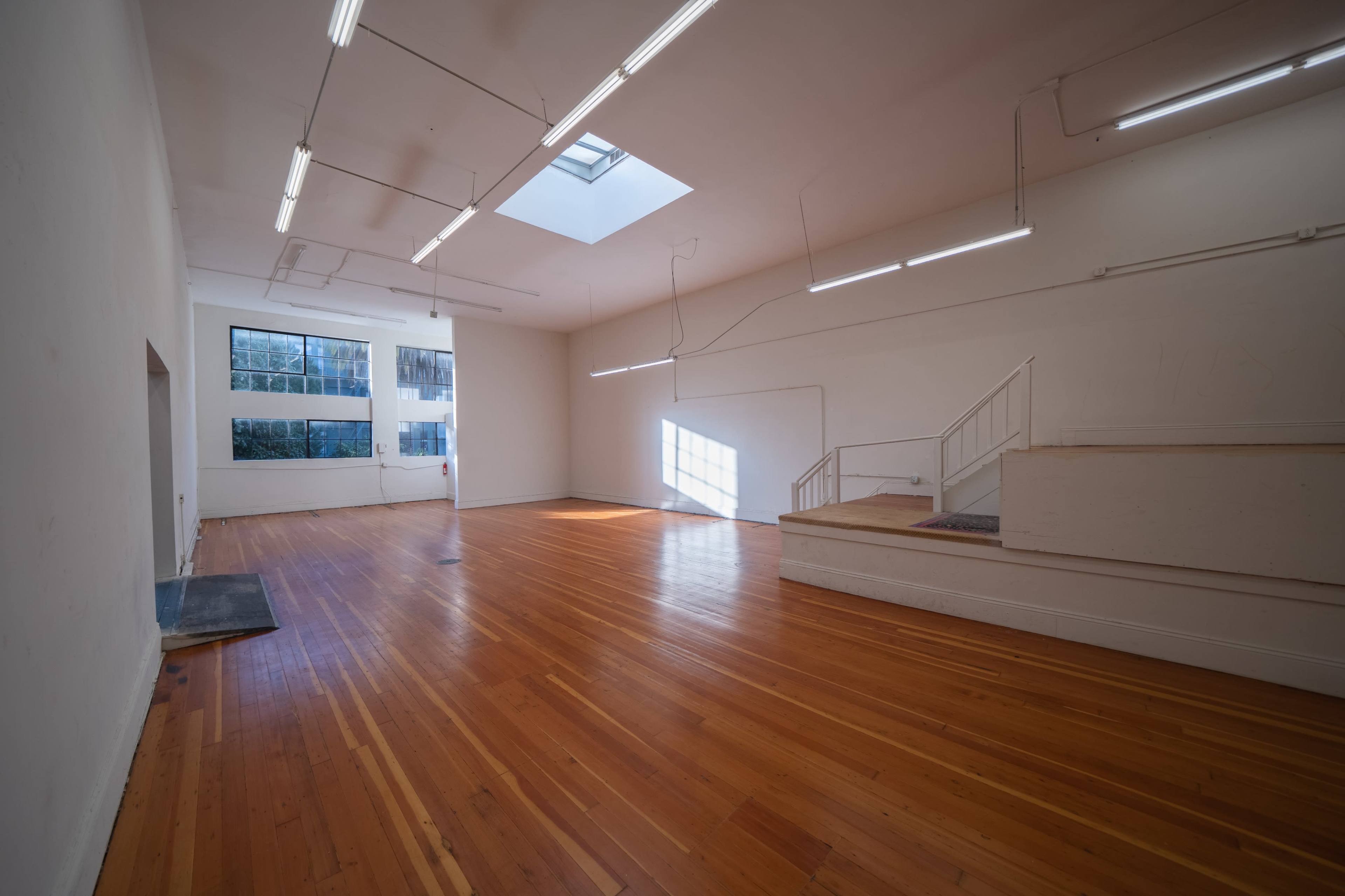 A spacious, empty room with hardwood floors, large windows, and fluorescent lighting.