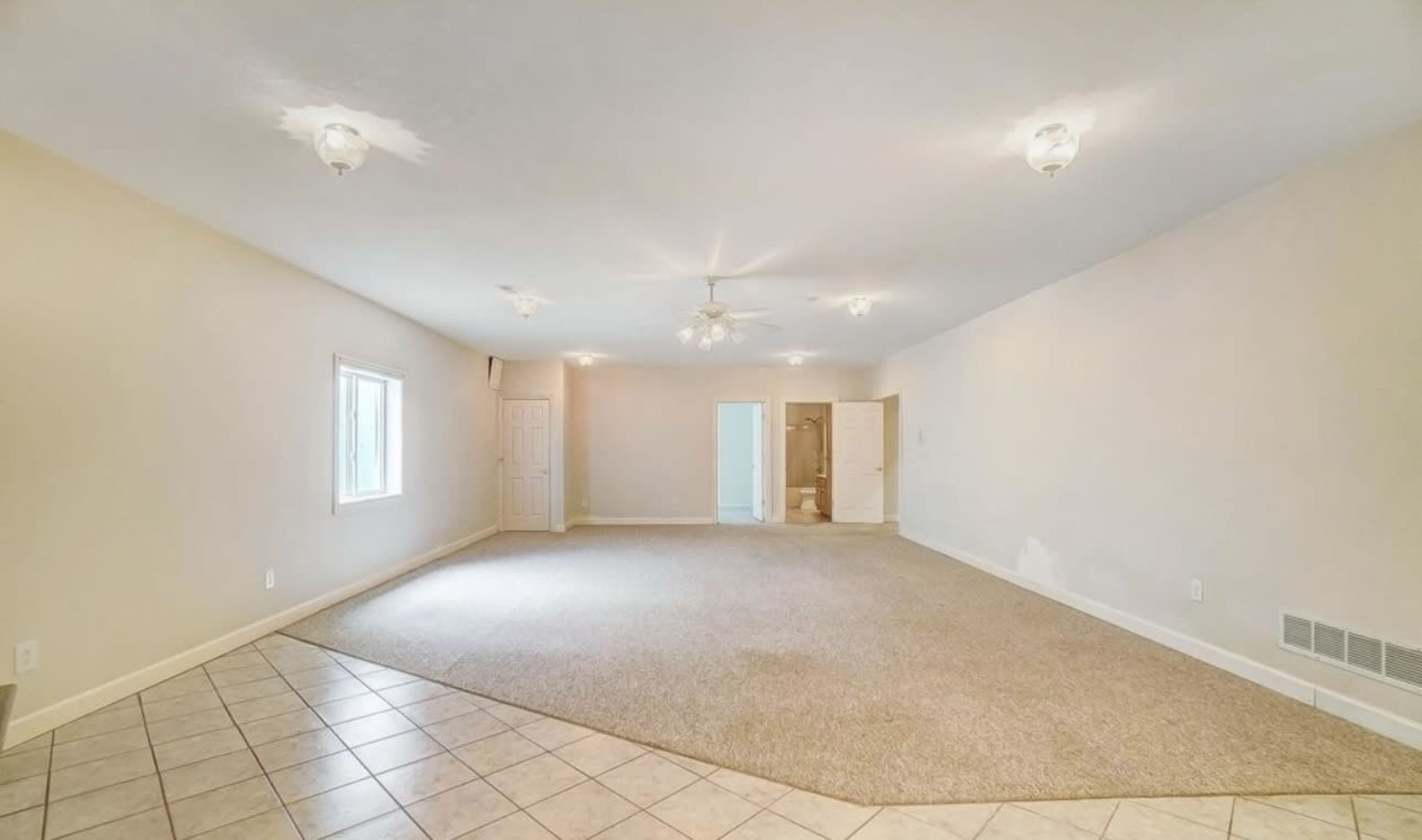 The image shows a spacious room with carpet flooring, a ceiling fan, and a door leading to a bathroom area in the background.