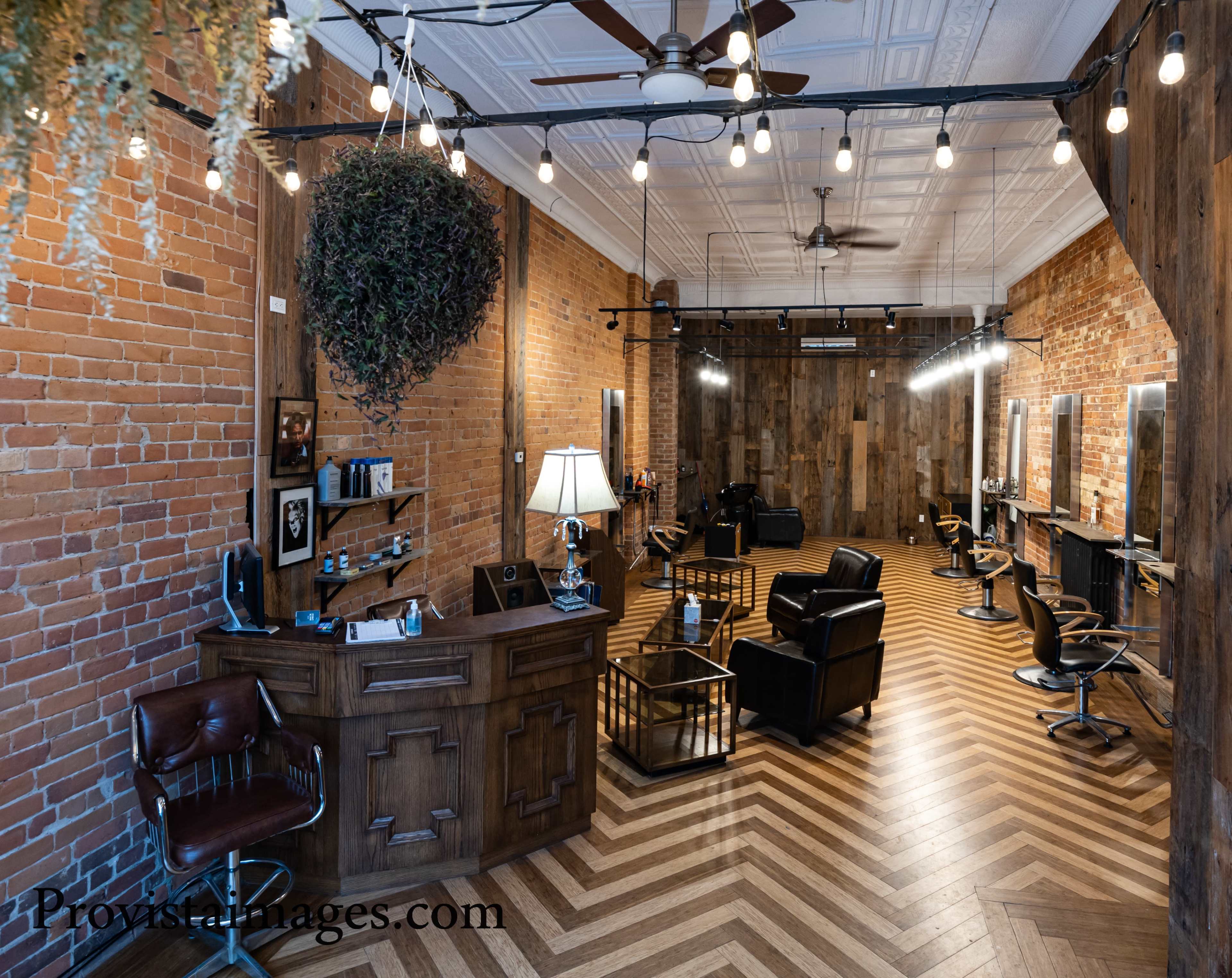 A spacious hair salon featuring brick walls, wooden accents, and a chevron-patterned floor with black salon chairs and illuminated mirrors.
