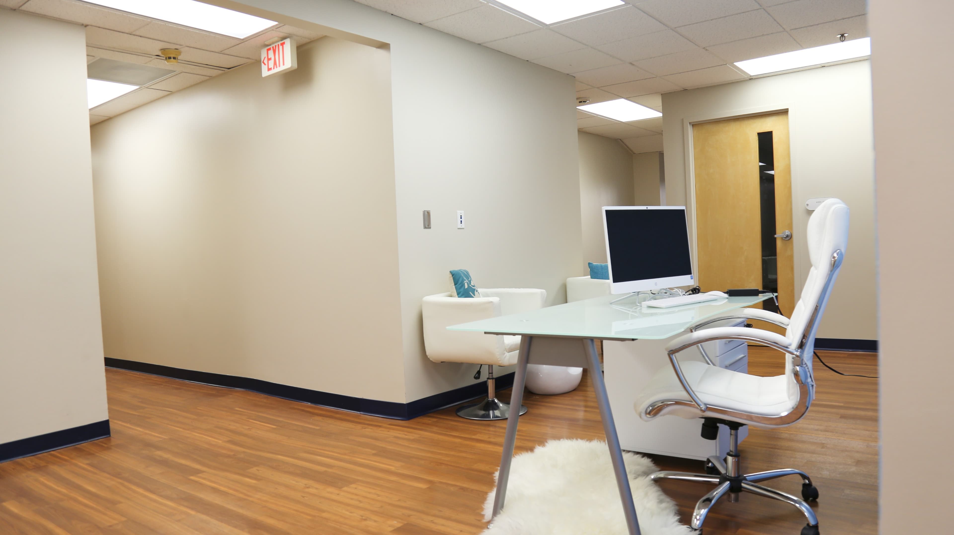 A modern office space features a white desk and chair with a computer, surrounded by light-colored walls and wooden flooring.
