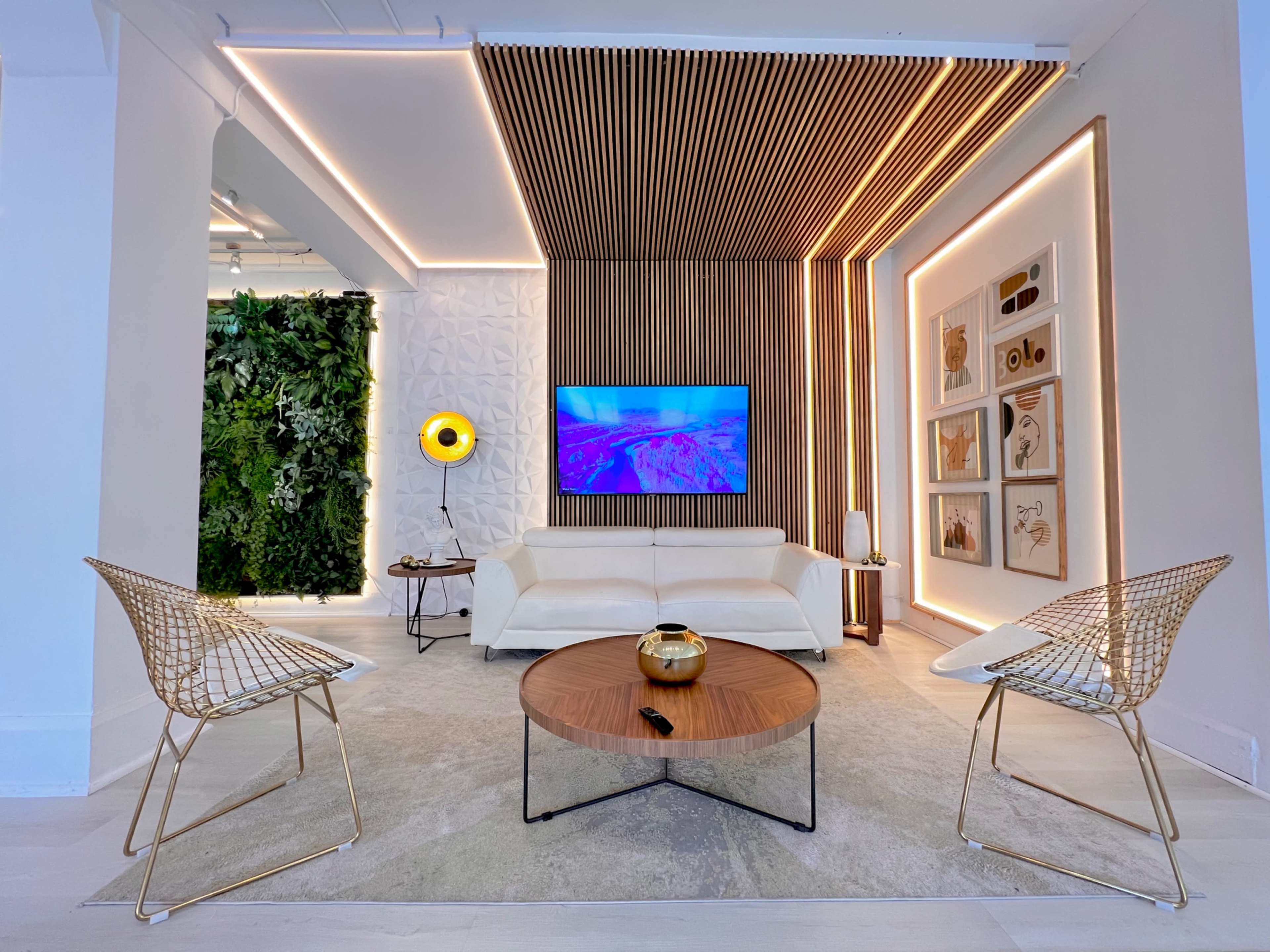 A modern living room features a white sofa, two wire chairs, a round coffee table, and a mounted television, with decorative wall accents and greenery.