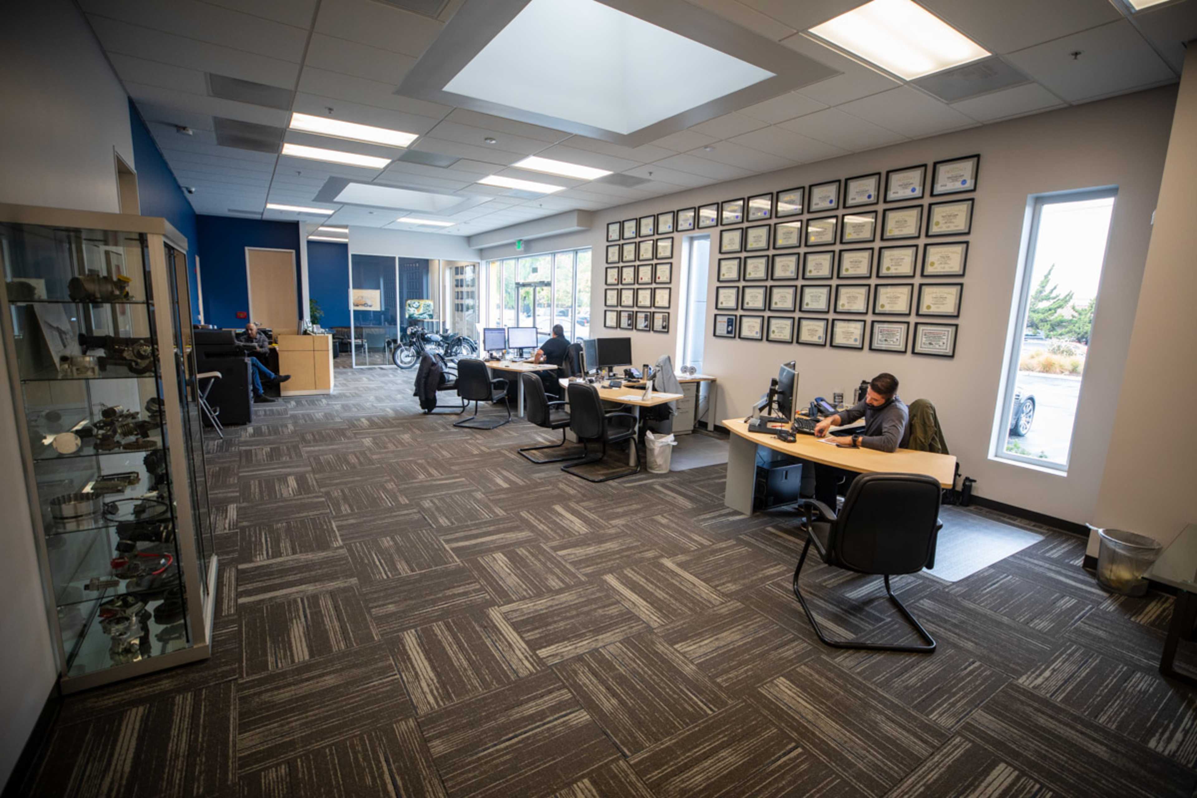 An office space with multiple workstations, a wall adorned with framed certificates, and people working at desks.