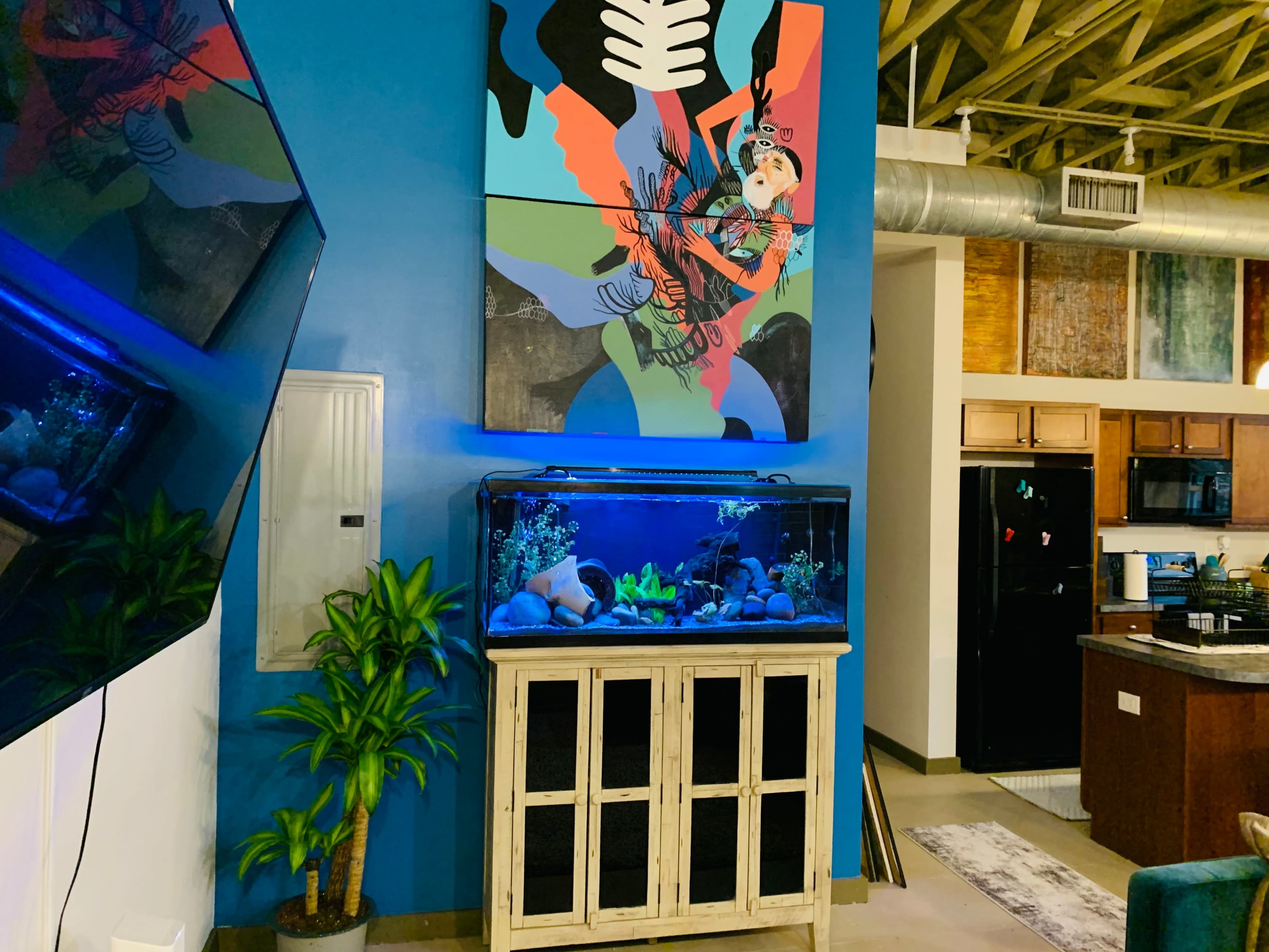 The image shows a modern interior with a large aquarium on a wooden stand, complemented by a colorful abstract painting on the wall, a potted plant nearby, and a kitchen area in the background.