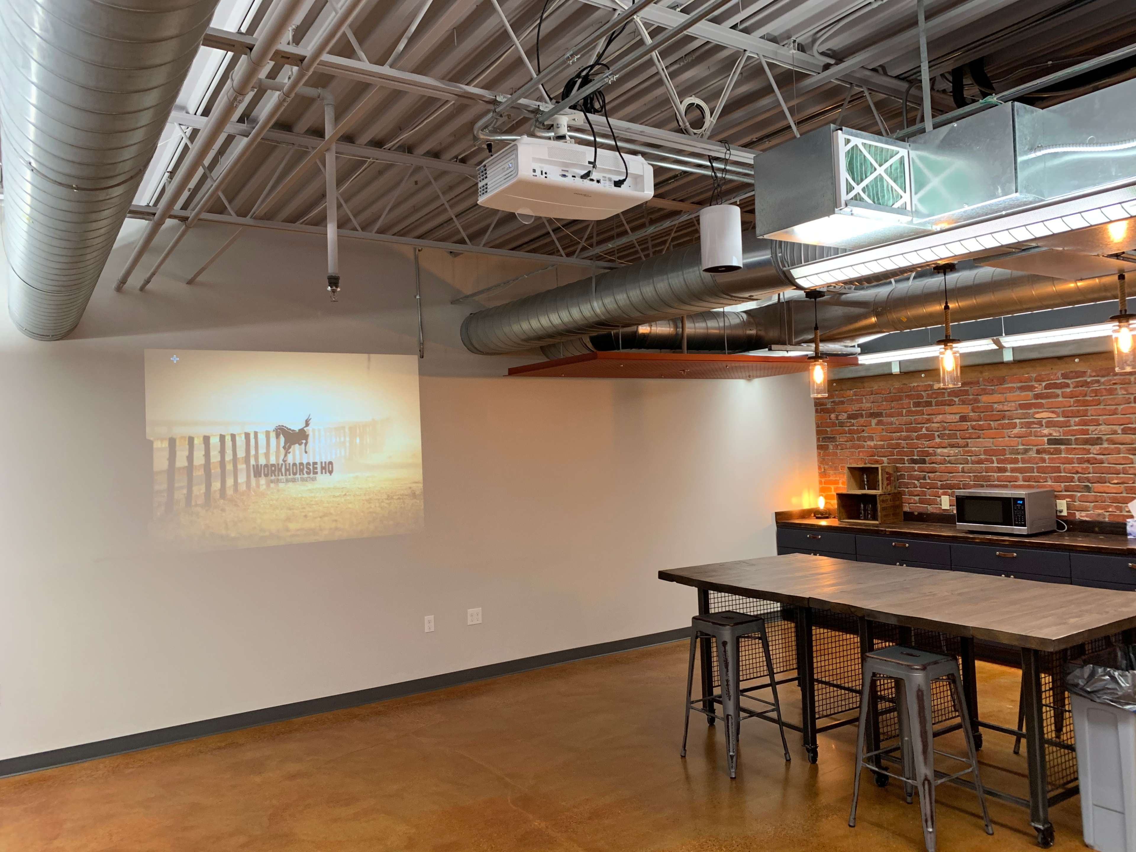 The image shows a modern kitchen space with a large wooden table, metal stools, and a projector displaying an image on the wall, set against a backdrop of brick and metal fixtures.
