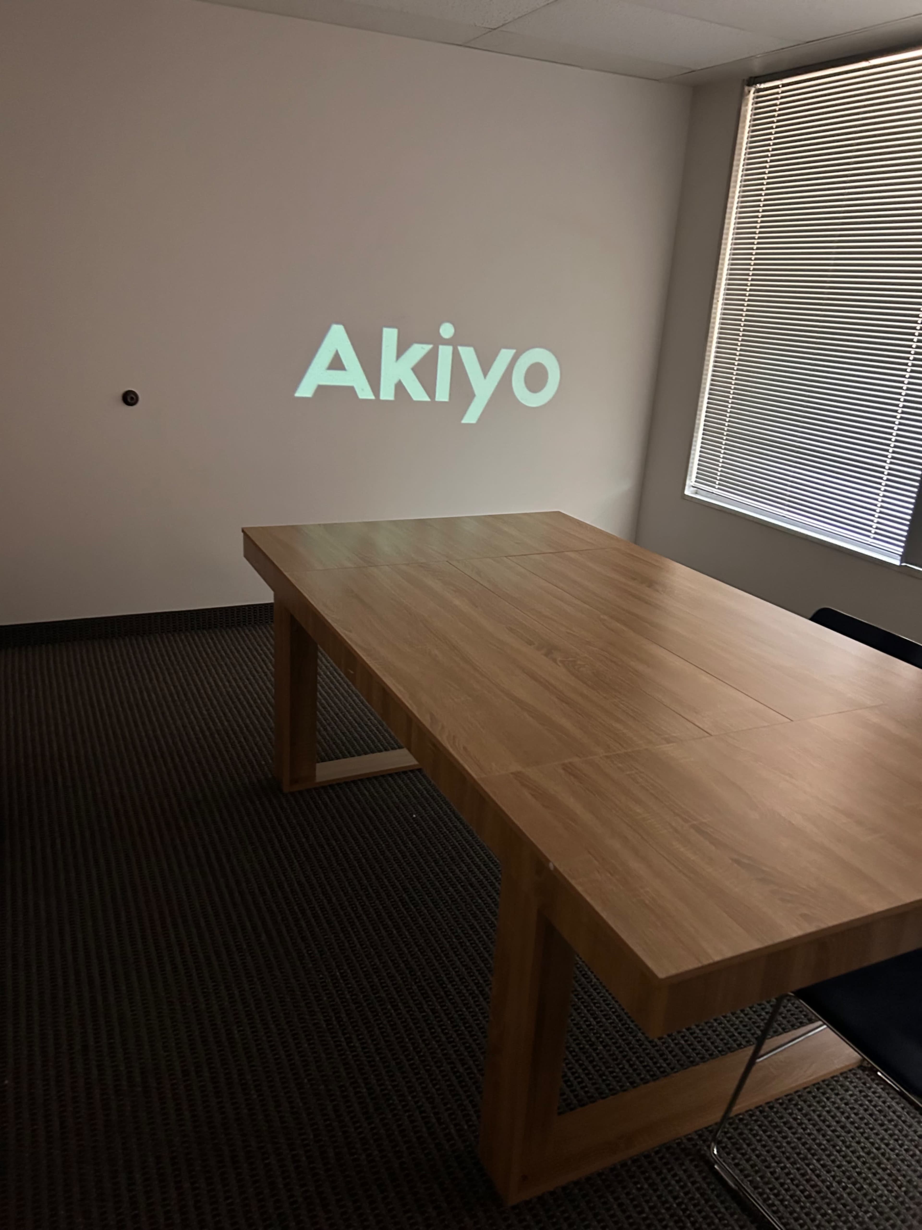 A wooden conference table is set in a well-lit room with the word "Akiyo" projected onto a blank wall.