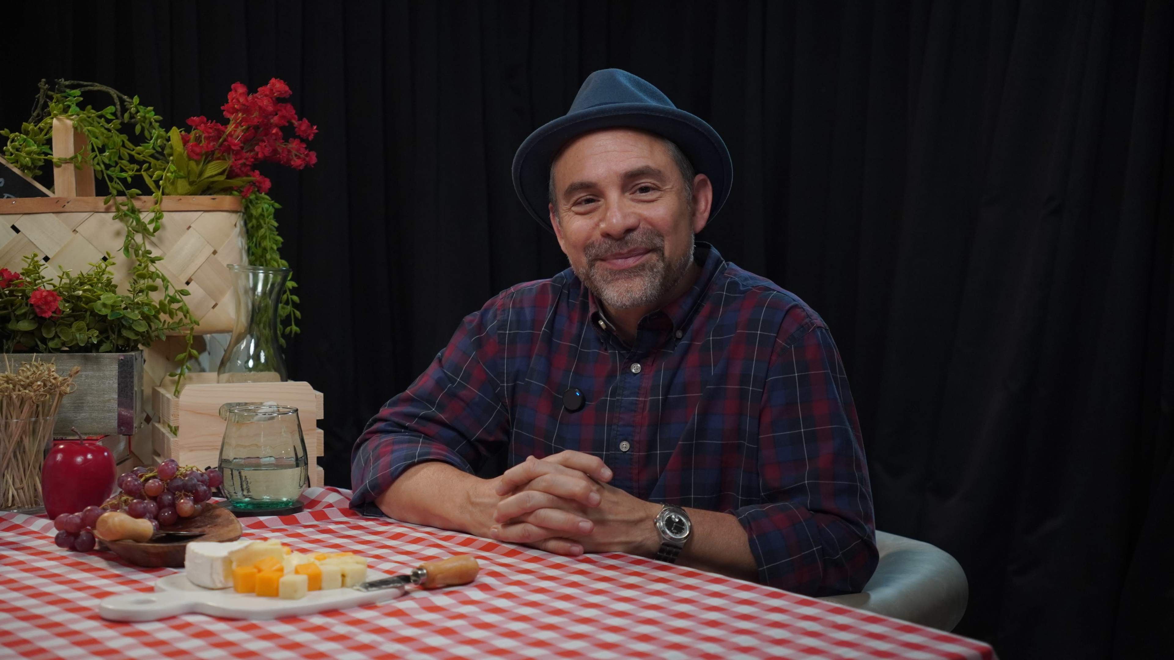 A man wearing a plaid shirt and a blue hat sits at a table with a checkered tablecloth, surrounded by food items like cheese, grapes, and a water pitcher.