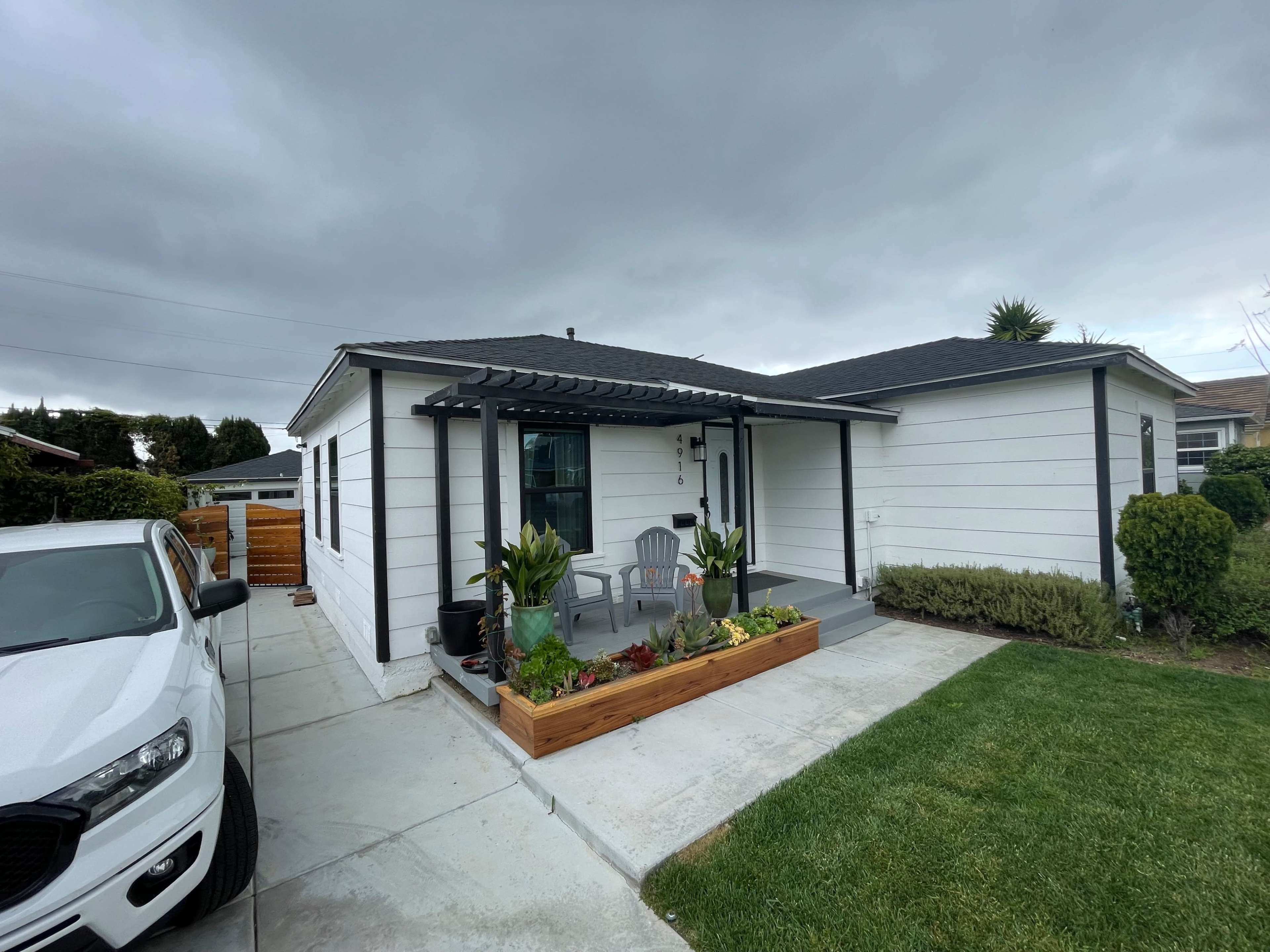 A single-story white house with a front porch and a flowerbed, surrounded by green grass and cloudy skies.