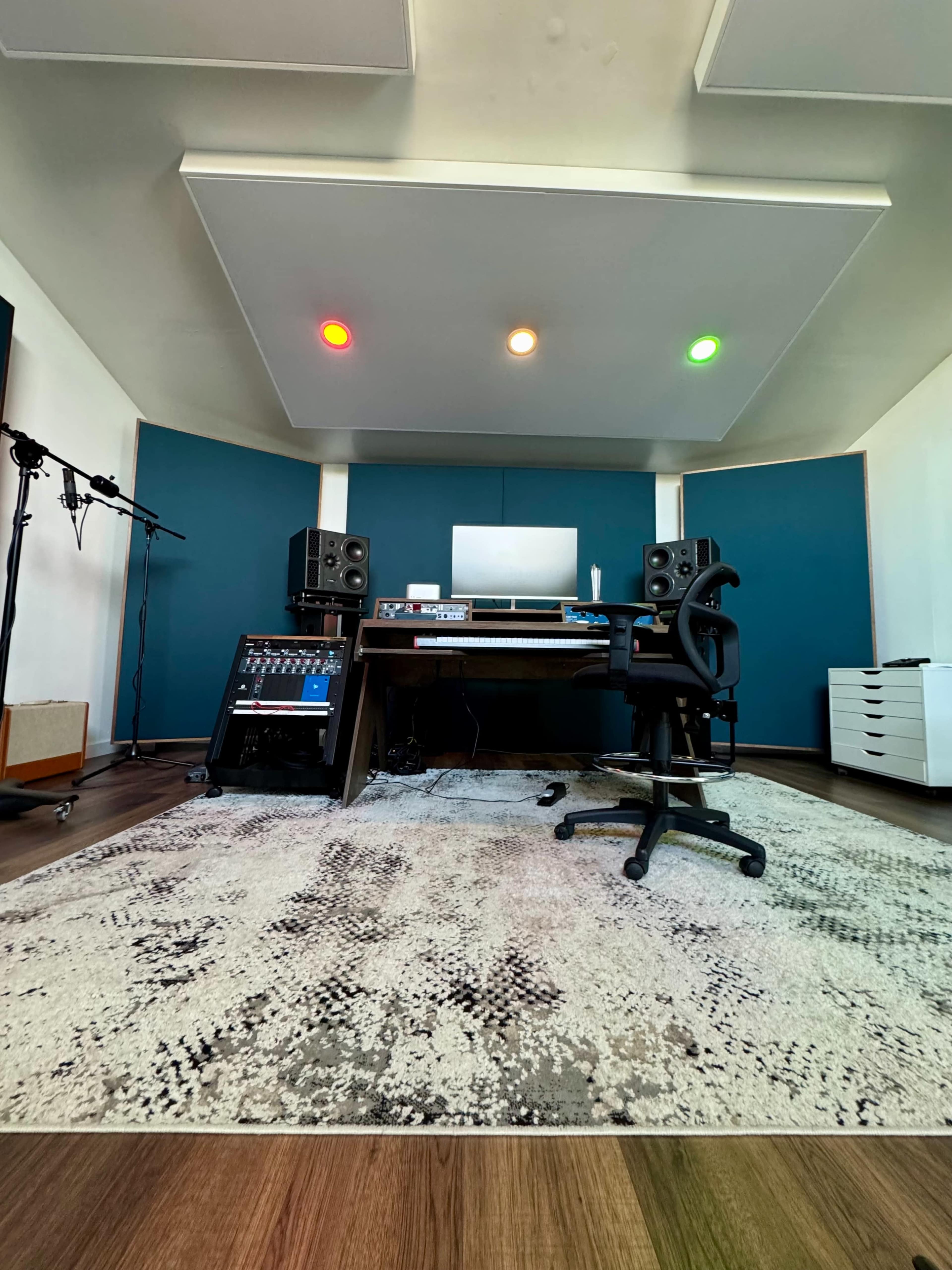 The image shows a modern audio recording studio with a central desk, audio monitors, and a patterned rug on the floor.