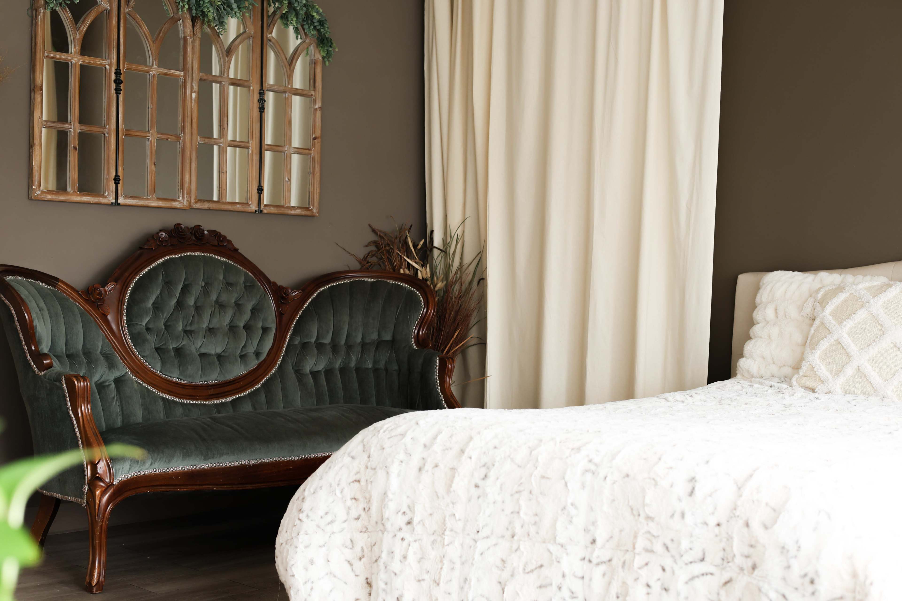 The image shows a cozy bedroom featuring a green velvet settee, a white bed with a textured blanket, and a large window draped with a cream-colored curtain.