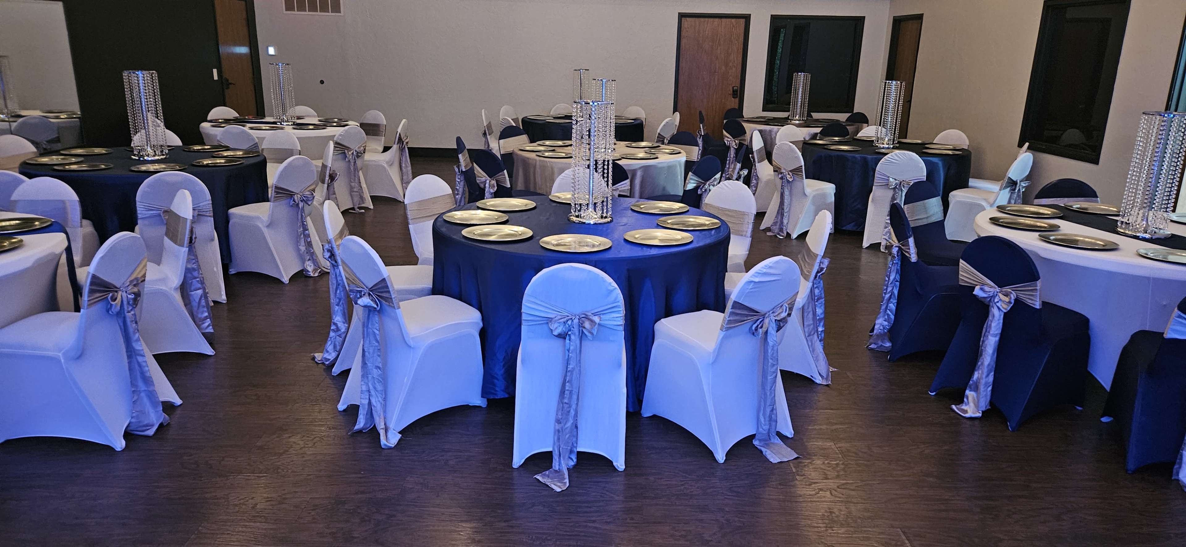 The image shows a banquet hall set up with round tables covered in navy and white tablecloths, each adorned with elegant centerpieces and chairs draped in white with blue bows.