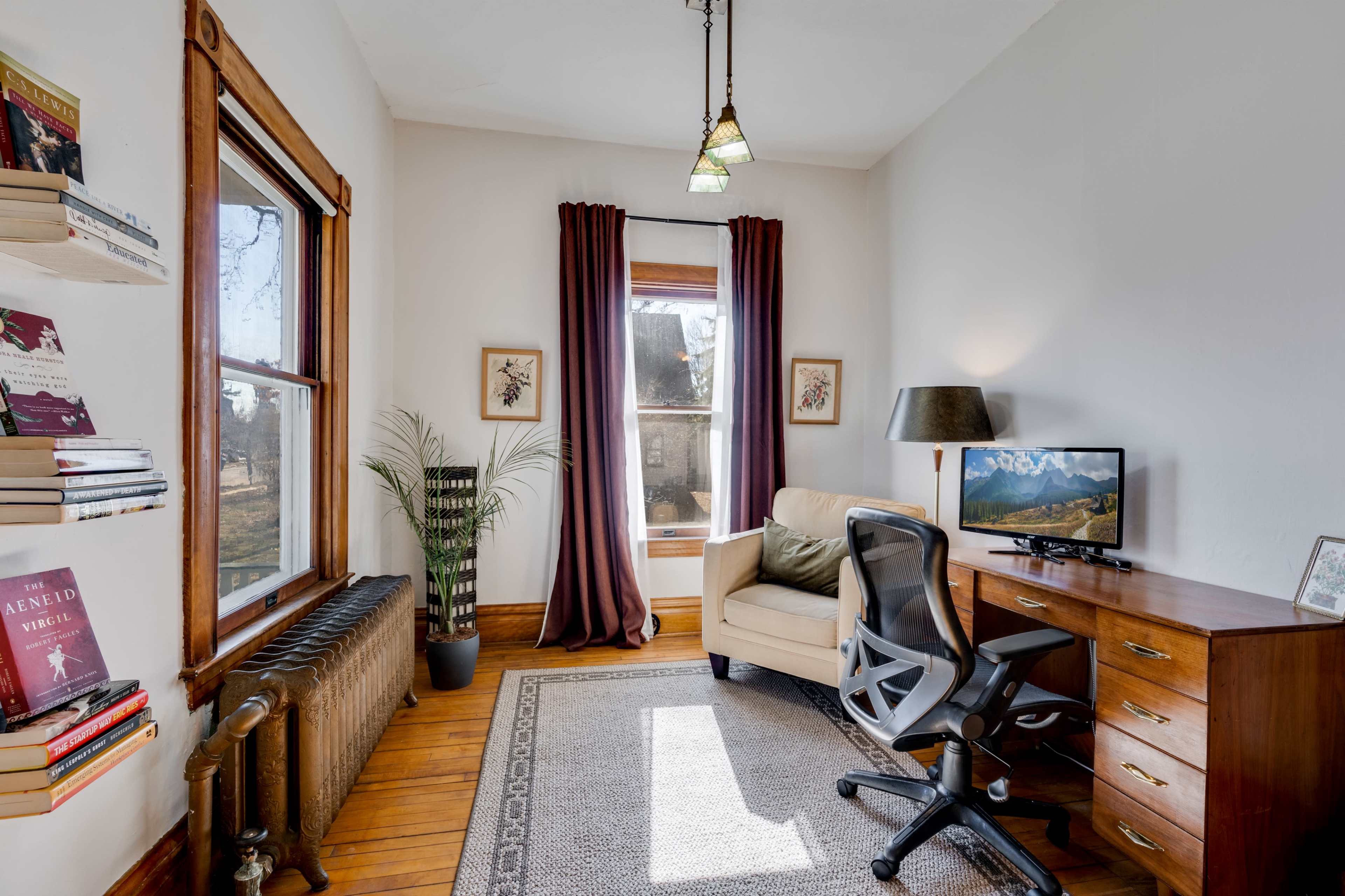 A cozy office features a desk with a monitor, a comfortable chair, and a large window framed by curtains, alongside a radiator and a bookshelf filled with books.