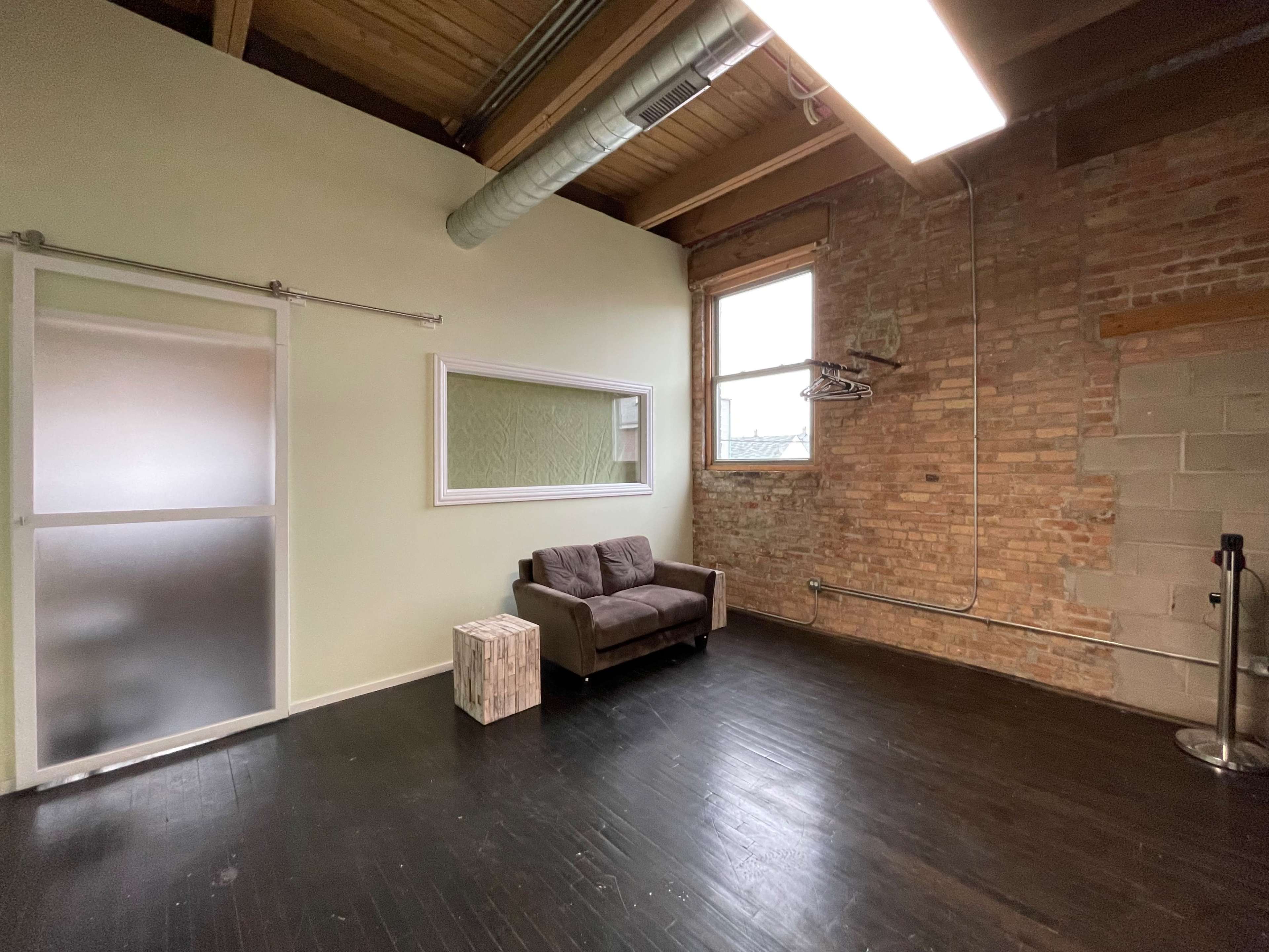 The image shows a spacious room with exposed brick walls, a dark wooden floor, a small brown couch, a mirror, and a frosted glass sliding door.