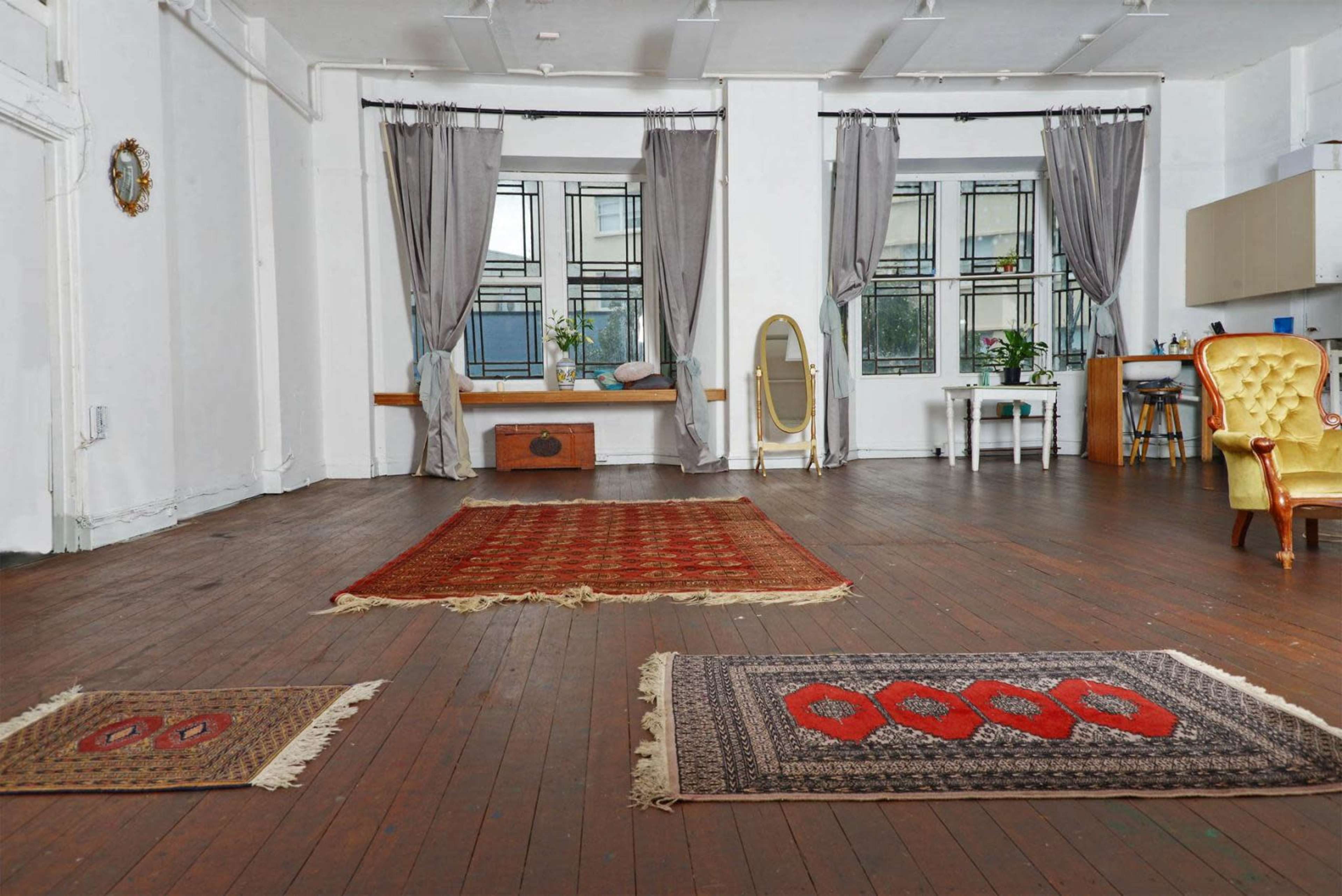 A spacious, well-lit room with wooden floors, two large windows dressed with curtains, and several rugs placed on the floor, along with a mirror and a small table in the background.