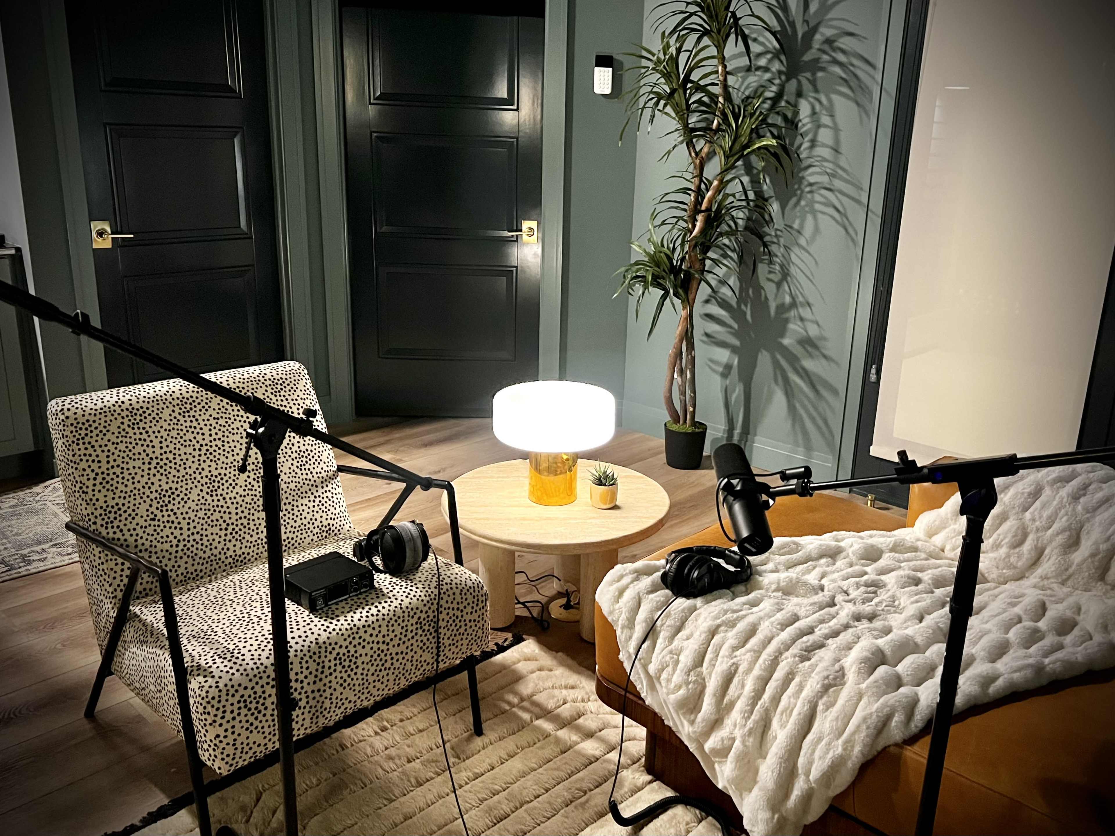 The image shows a cozy podcast recording setup with two chairs, a circular table containing a lamp and a small plant, and two microphones on stands.