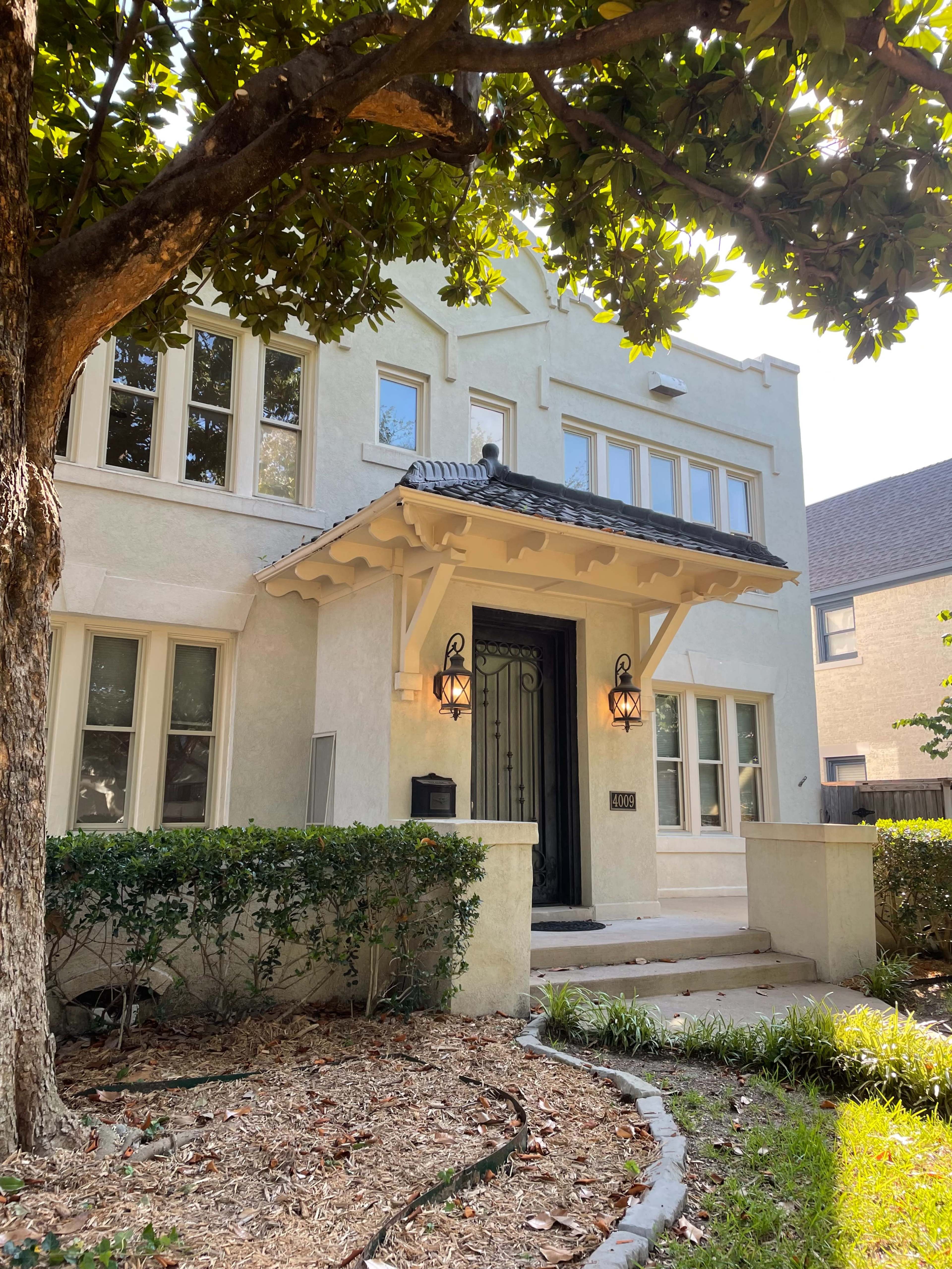 A two-story, light-colored house with decorative facade details, flanked by a garden and large tree.