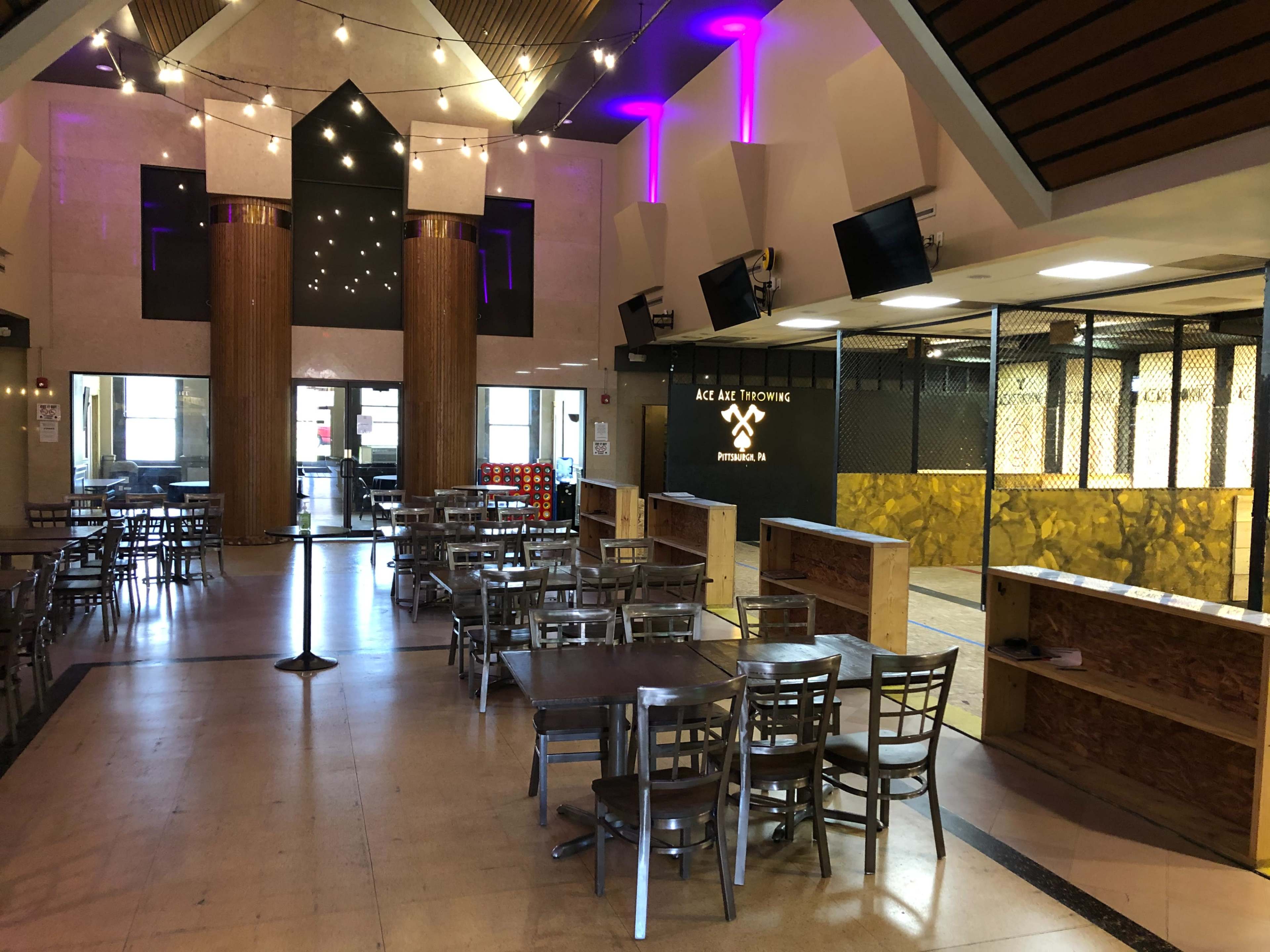 The interior of a venue with rows of metal tables and chairs, a reception area, and a space designed for activities, illuminated with purple lighting.