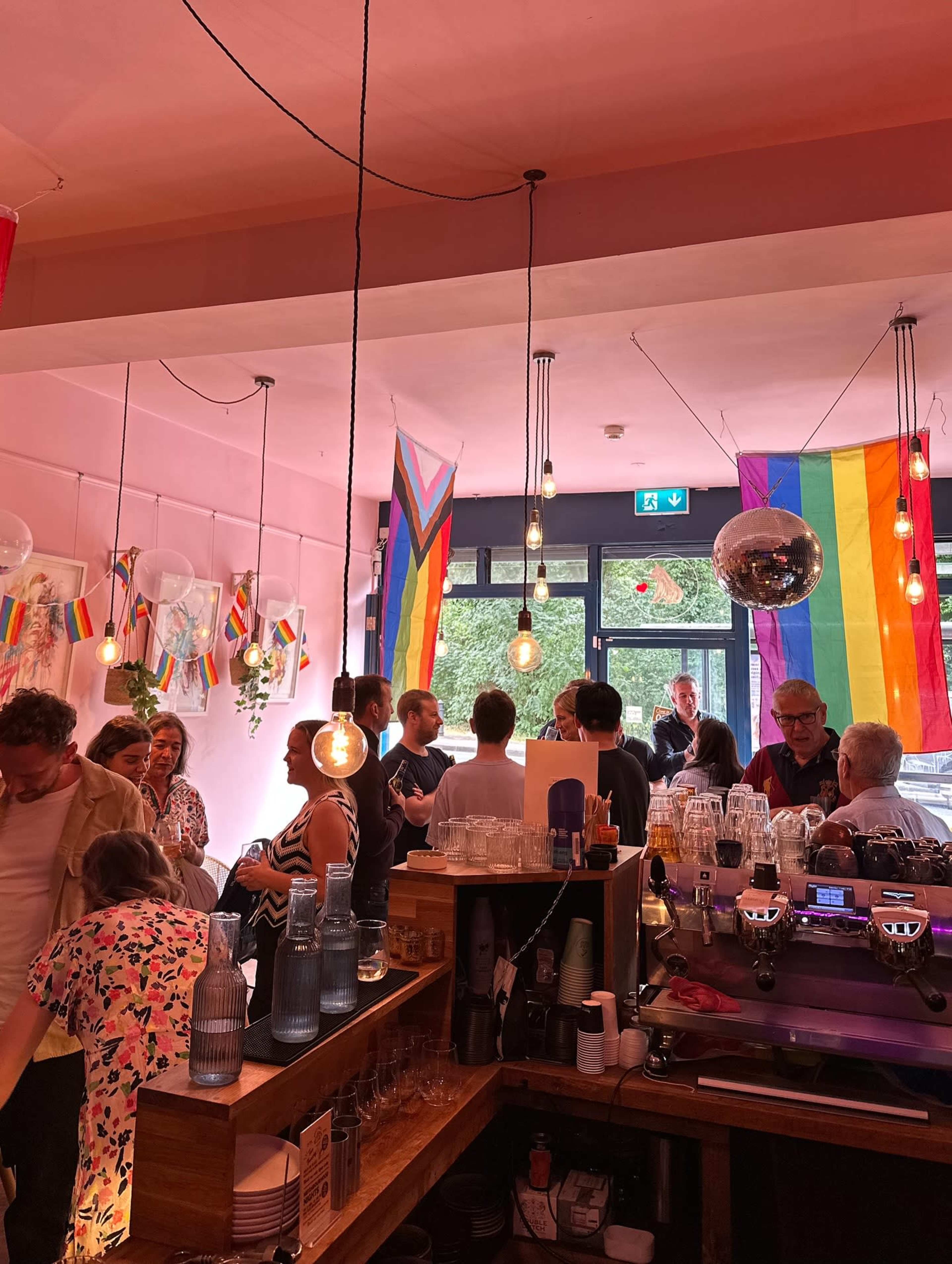 The image shows a vibrant bar interior decorated with various rainbow flags, where a crowd of people gathers around the bar area and socializes under warm hanging lights.