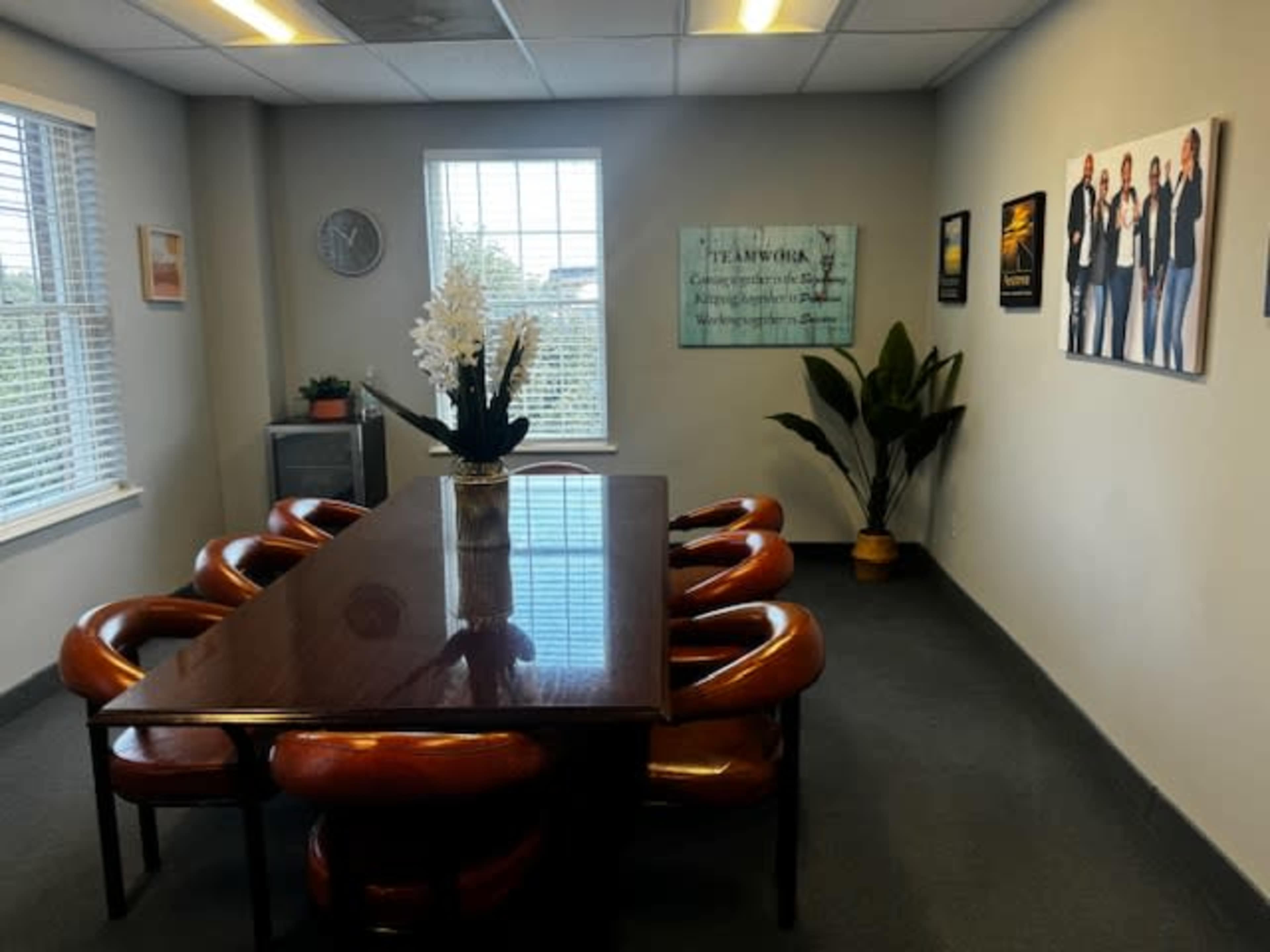 A conference room features a long table surrounded by brown chairs, with a large plant and decorative artwork on the walls.