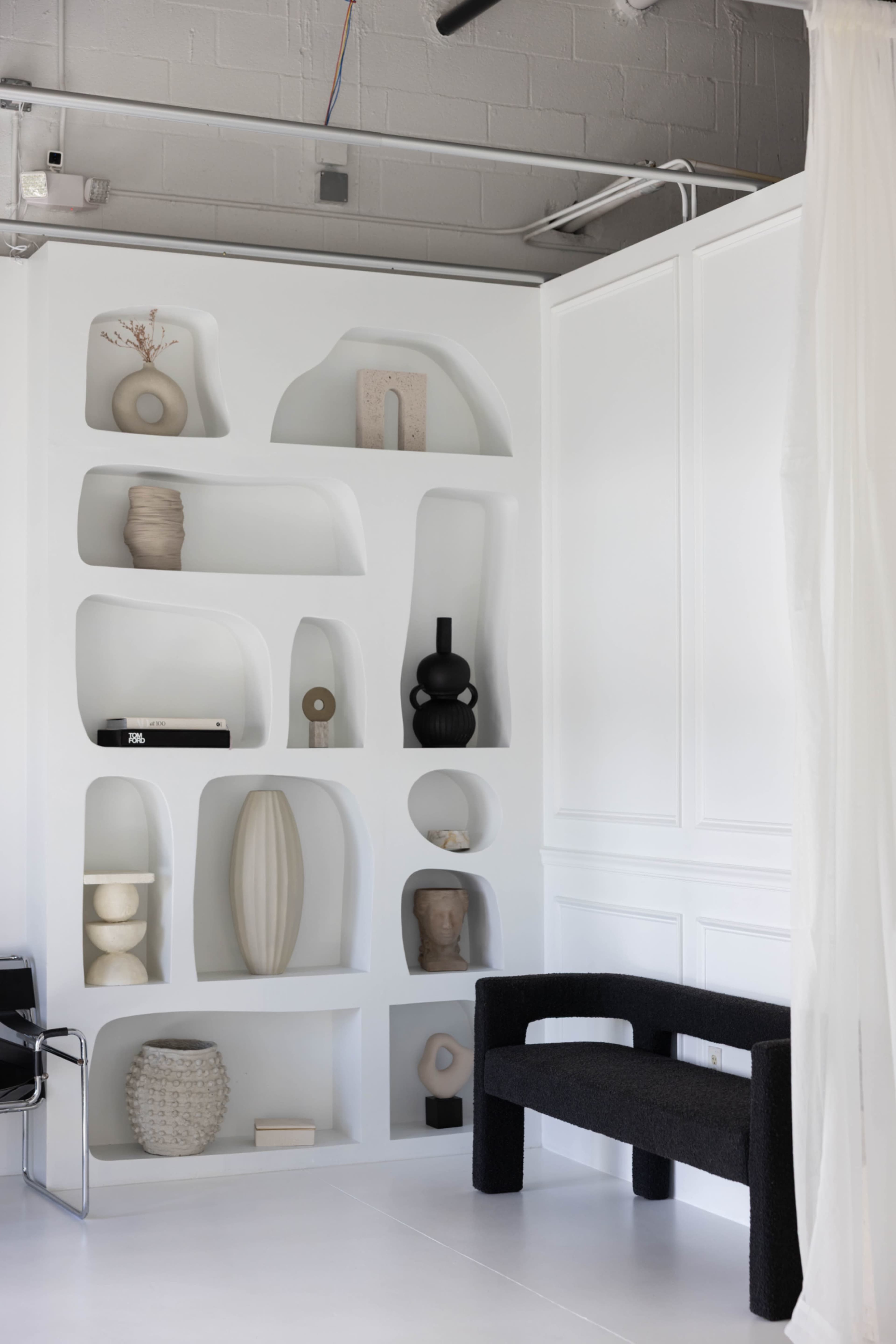 The image features a modern interior with a white wall displaying various sculptural objects and a black bench against it.