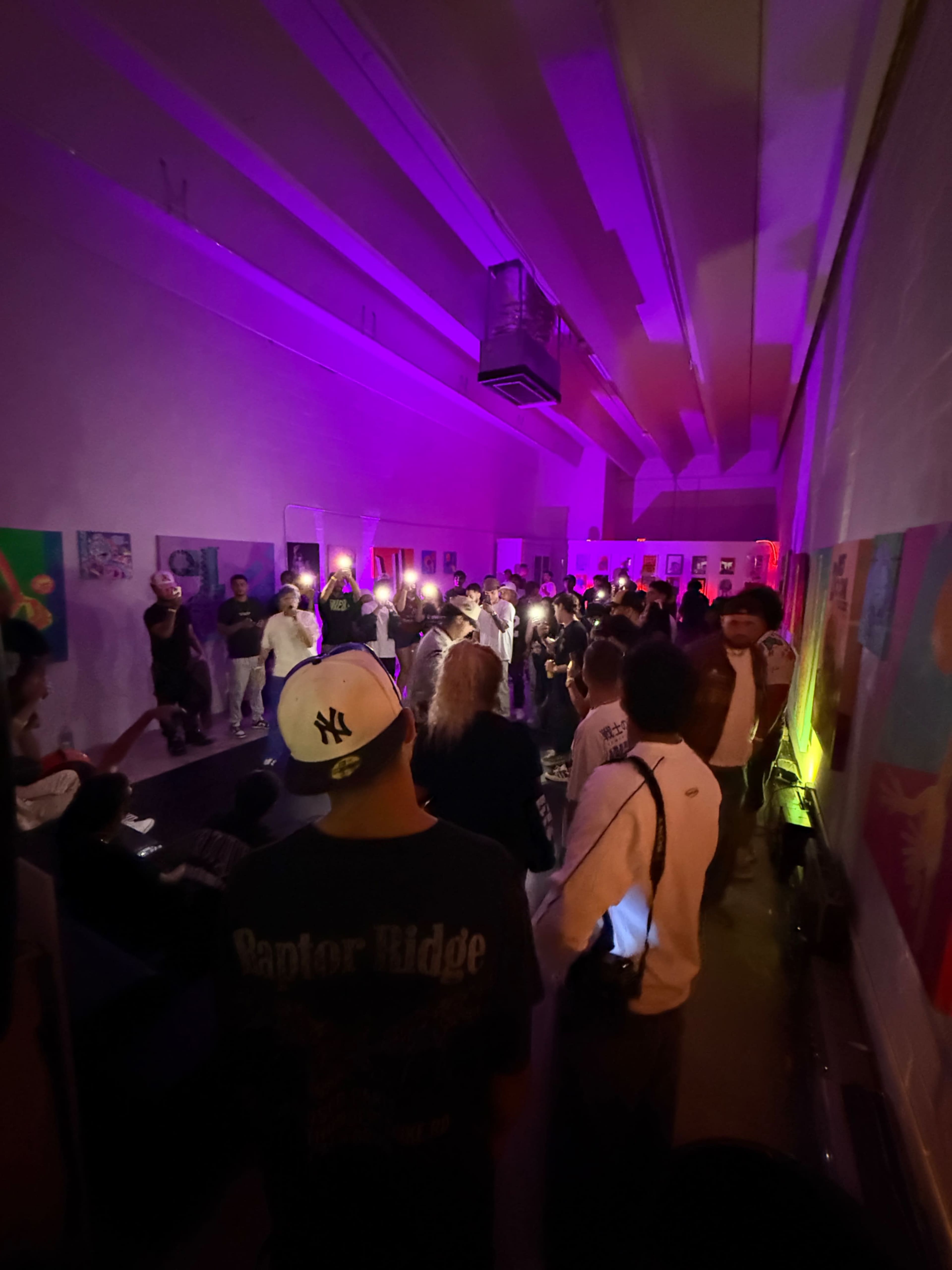 A large crowd gathers inside a gallery with colorful lighting and artwork on the walls.