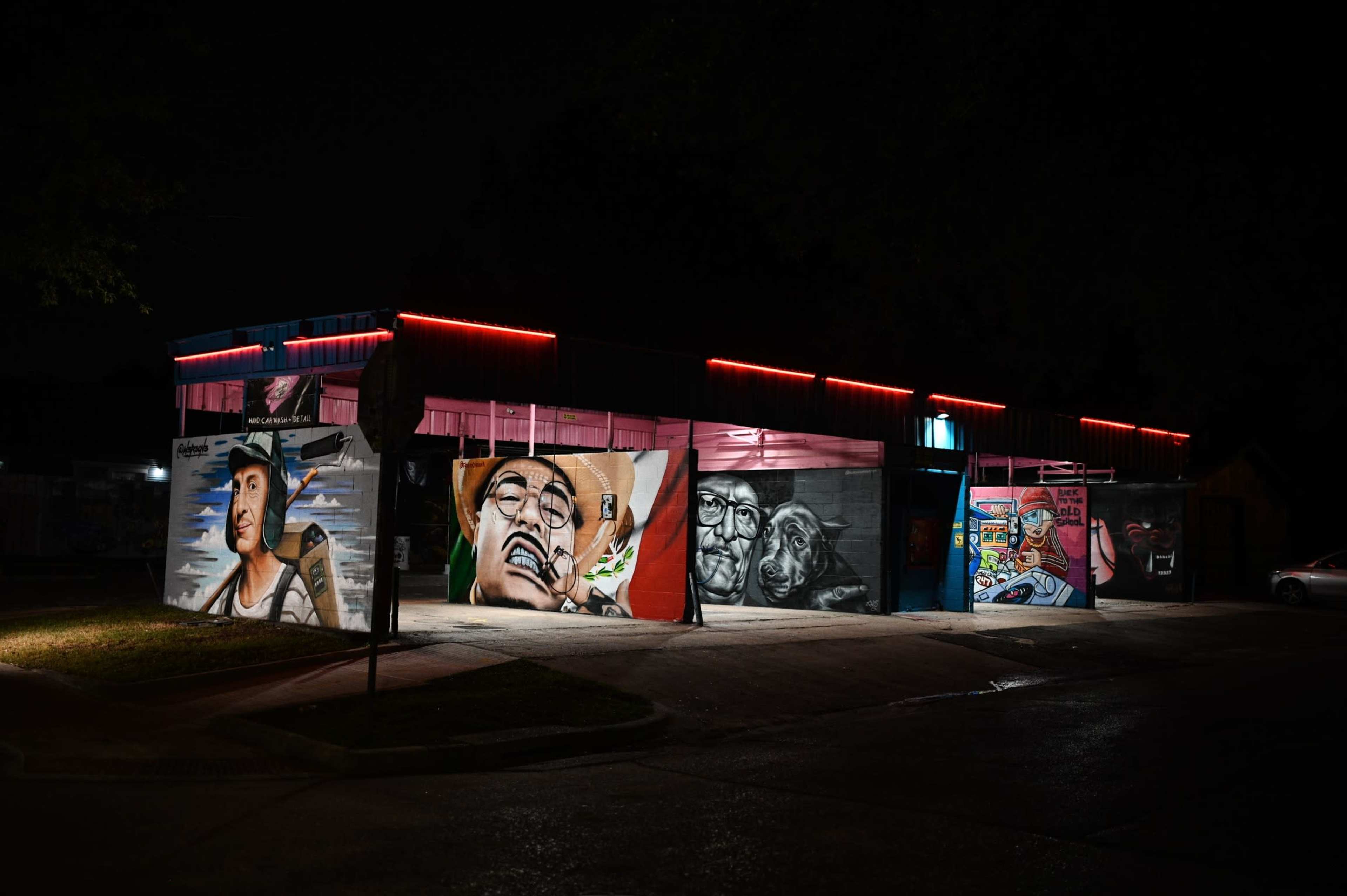 A building at night displays vibrant murals on its exterior walls, highlighted by neon red lights.
