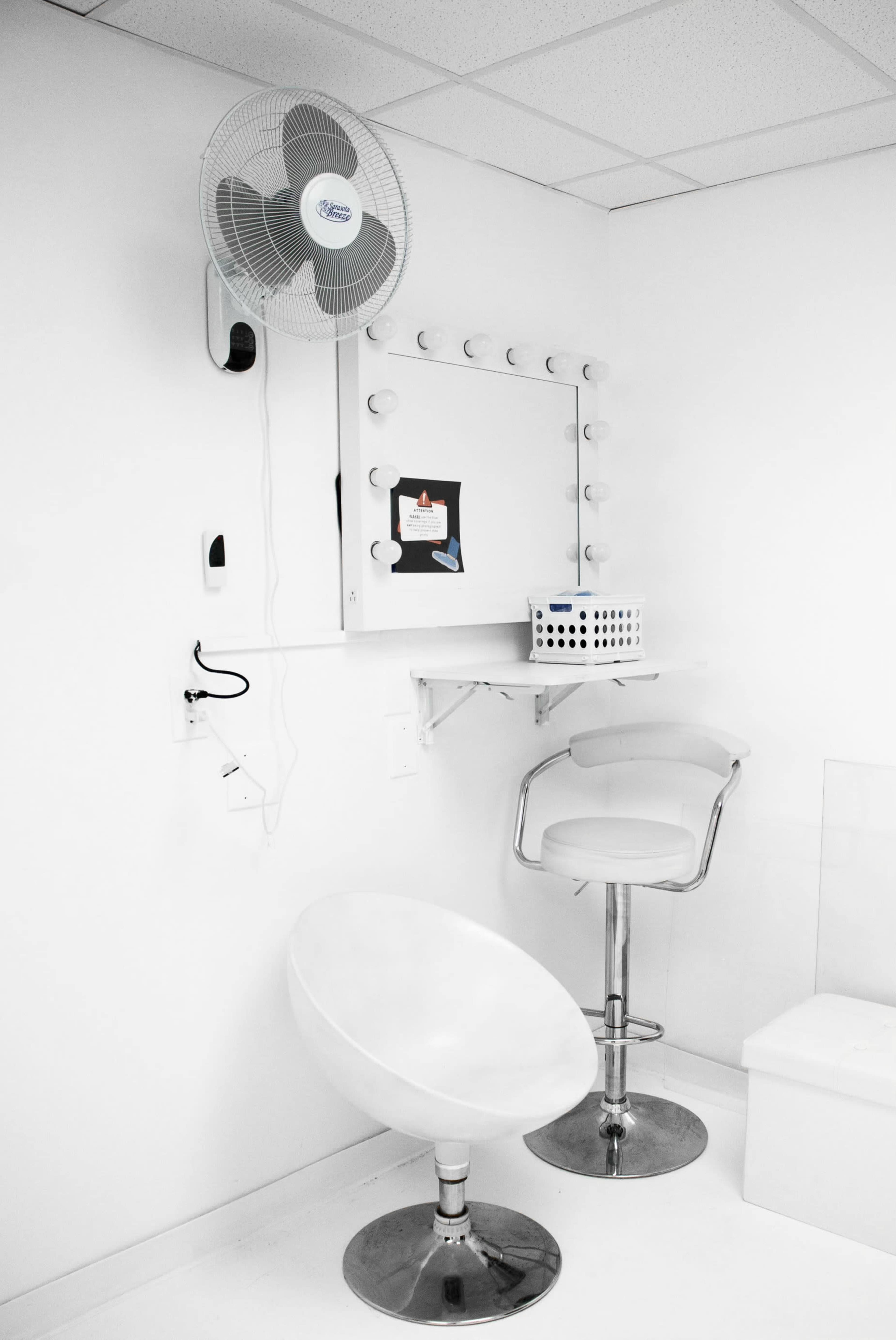 A minimalist dressing room featuring a circular makeup mirror with lights, a fan on the wall, a white stool, and a metallic barstool.
