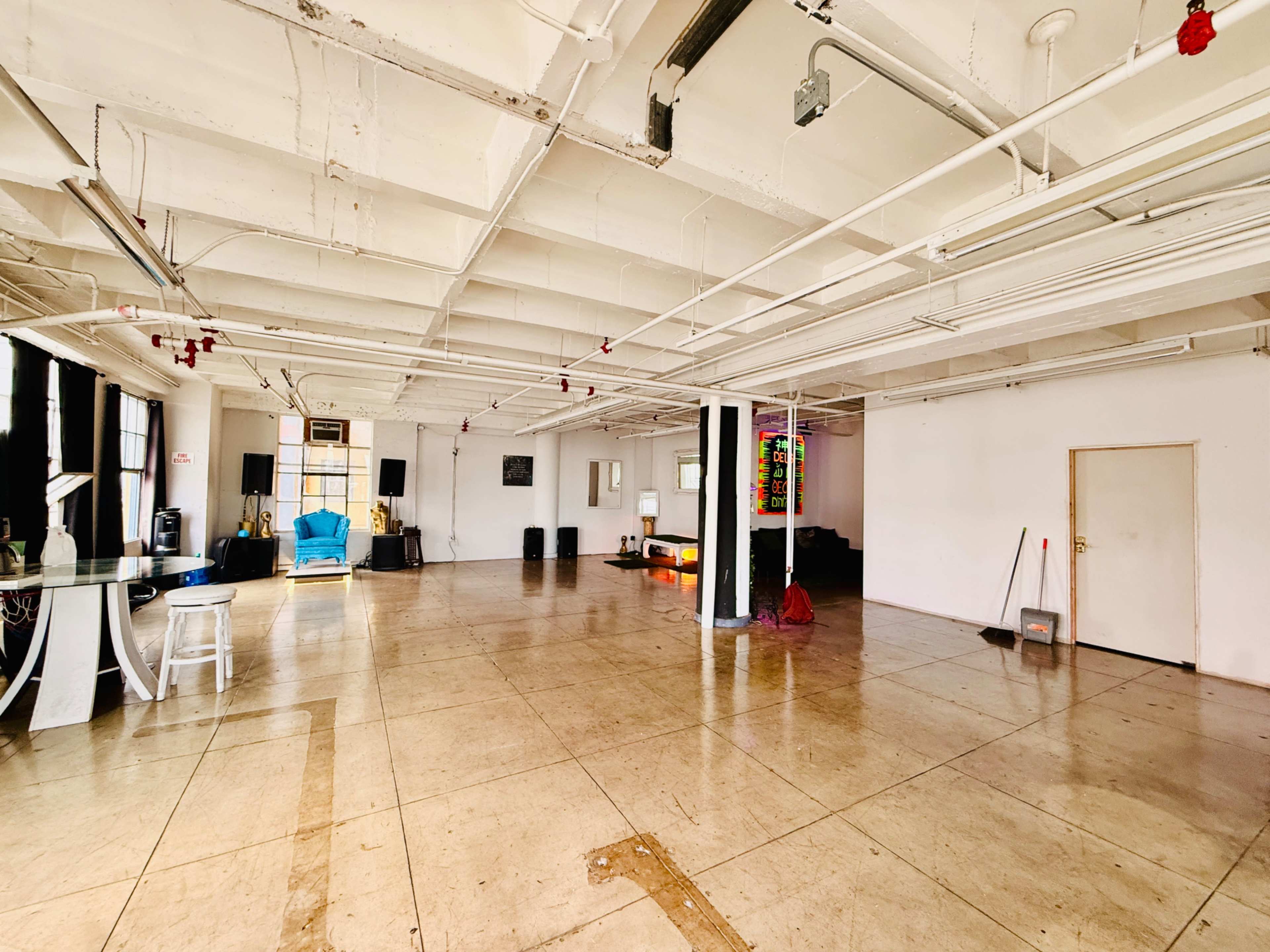 The image shows a spacious, empty room with concrete floors and exposed ceiling beams, featuring minimal furnishings and bright natural light.