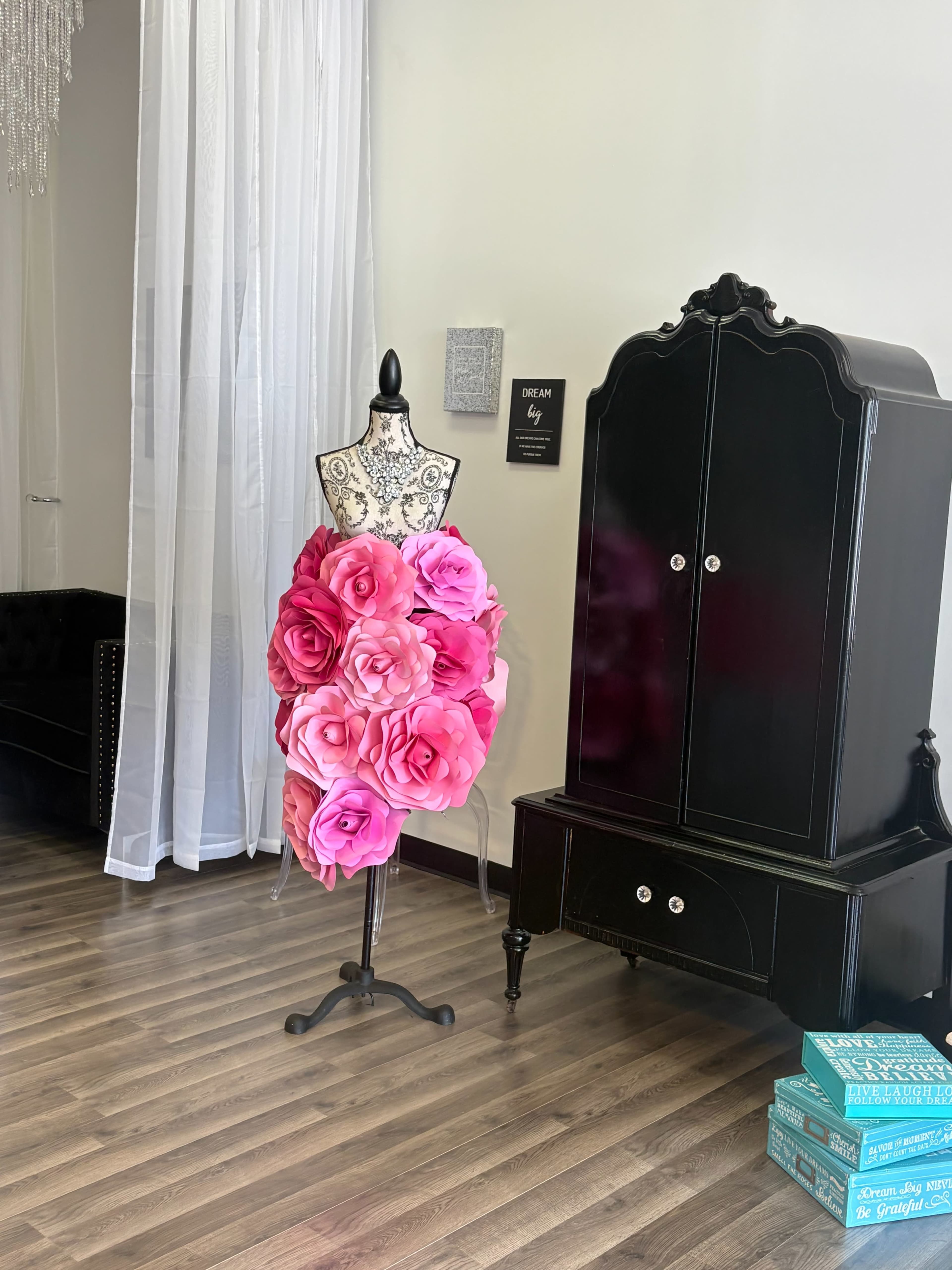 A dress form adorned with large pink paper flowers stands in a brightly lit room next to a black cabinet.