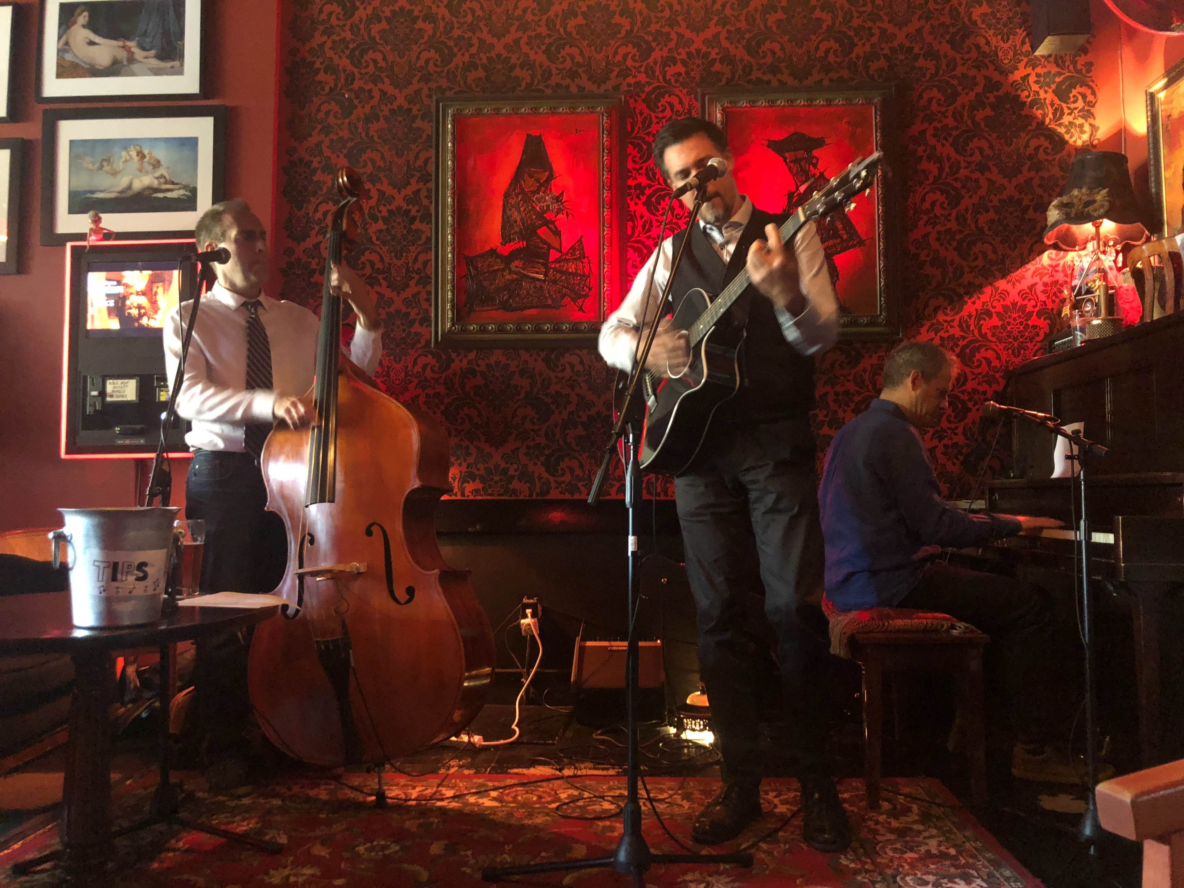Three musicians perform live music in a cozy venue, with one playing guitar and singing, another playing double bass, and the third on piano.