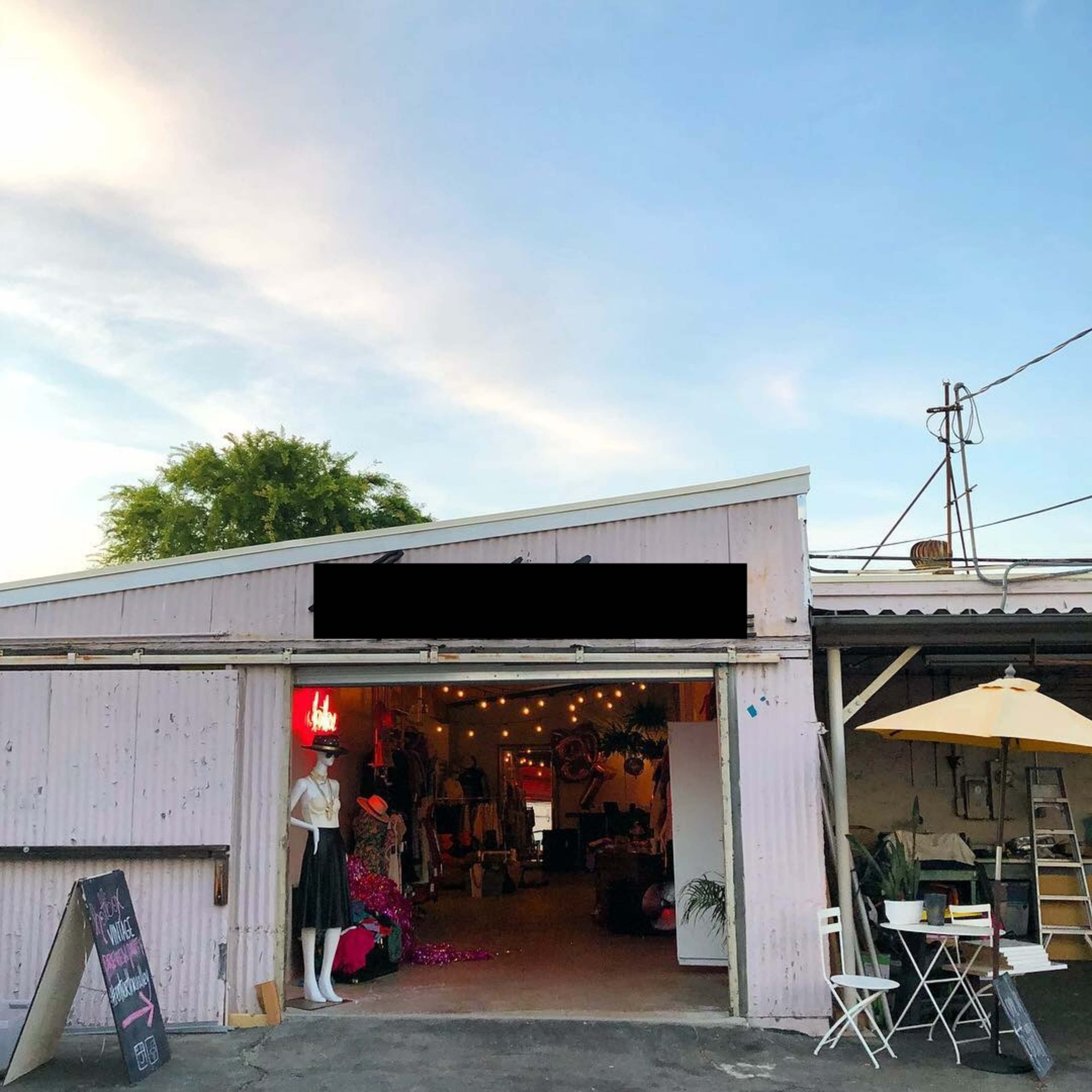 A pink metal building with an open door displays mannequins and items for sale, surrounded by outdoor seating under an umbrella.