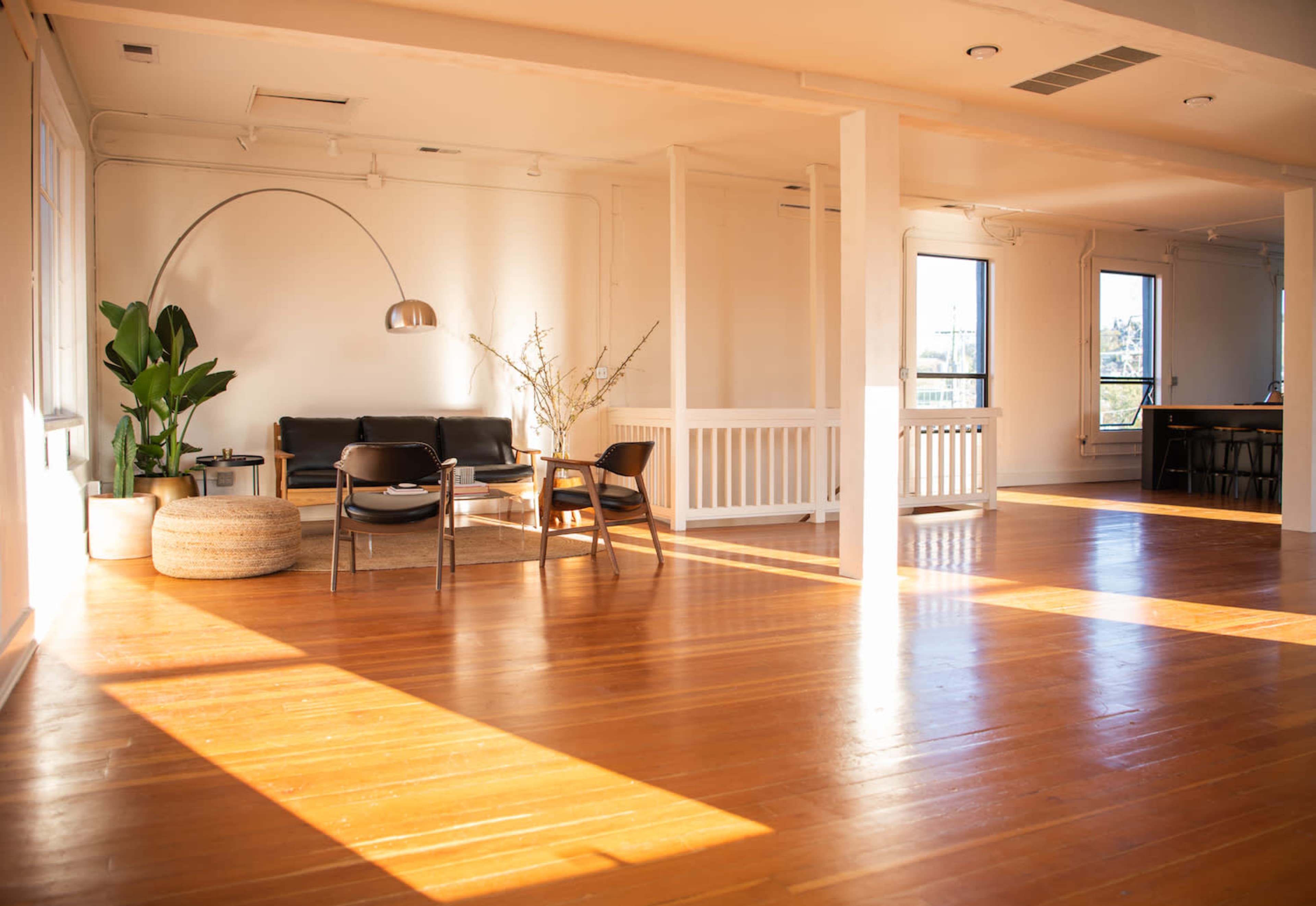 A spacious, well-lit room with polished wooden floors, featuring a black sofa, a large plant, a round pouf, and an arc floor lamp.
