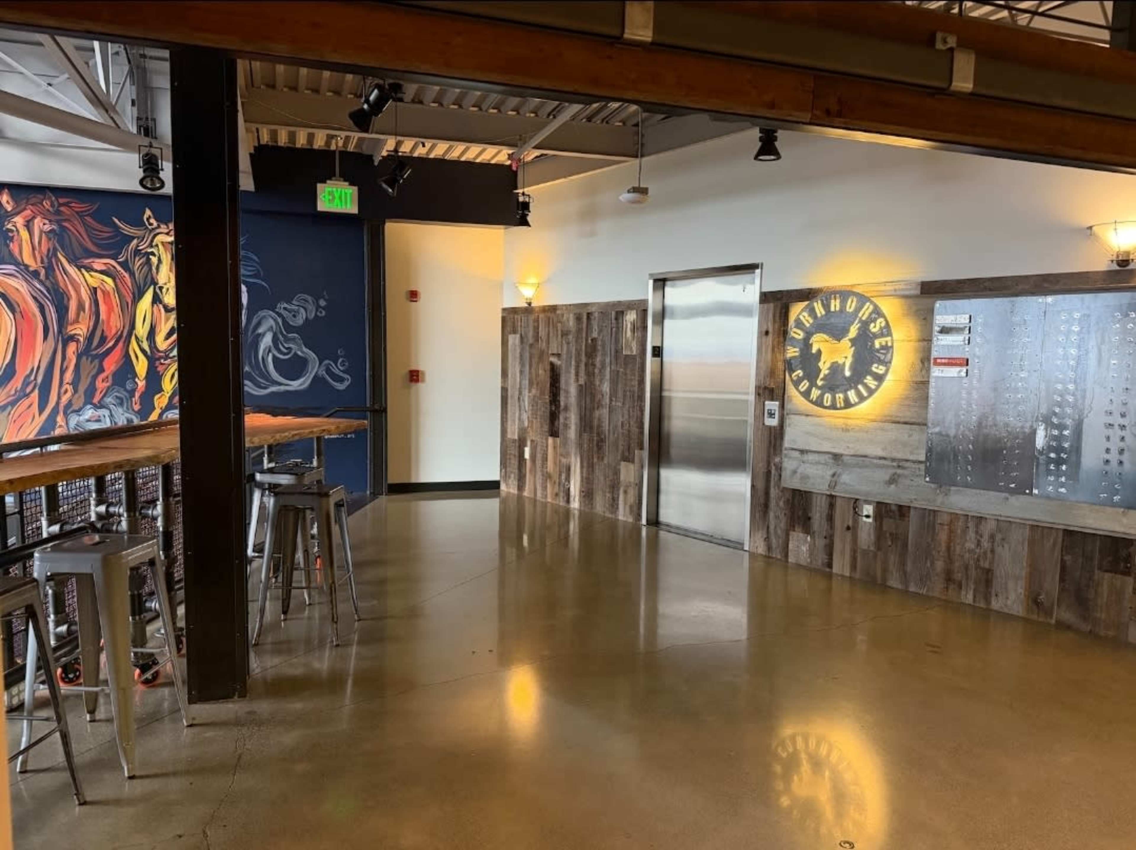 The image shows a modern, industrial-style interior of a restaurant or bar featuring a prominent mural, metal stools, and an elevator door.