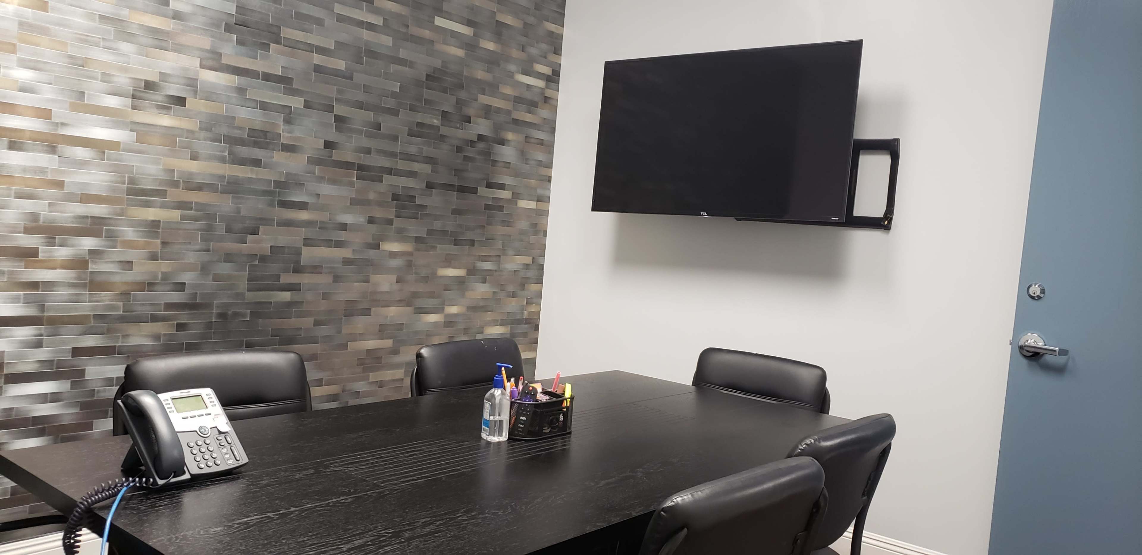 A conference room features a black table surrounded by six chairs, a wall-mounted television, and a stone-patterned accent wall.