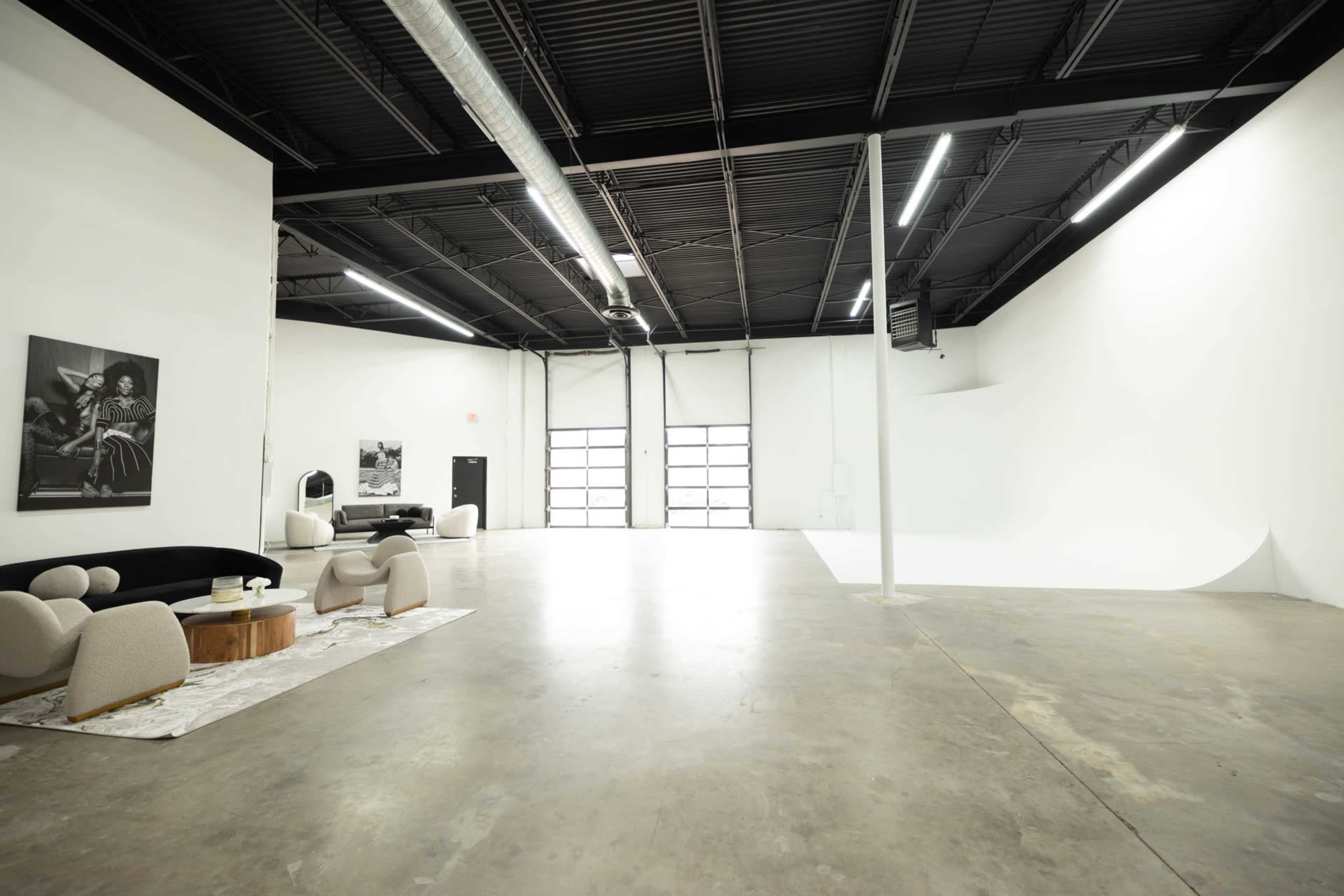 The image shows an empty studio space with a polished concrete floor, white walls, and a large backdrop area, furnished with a few modern chairs and a table.