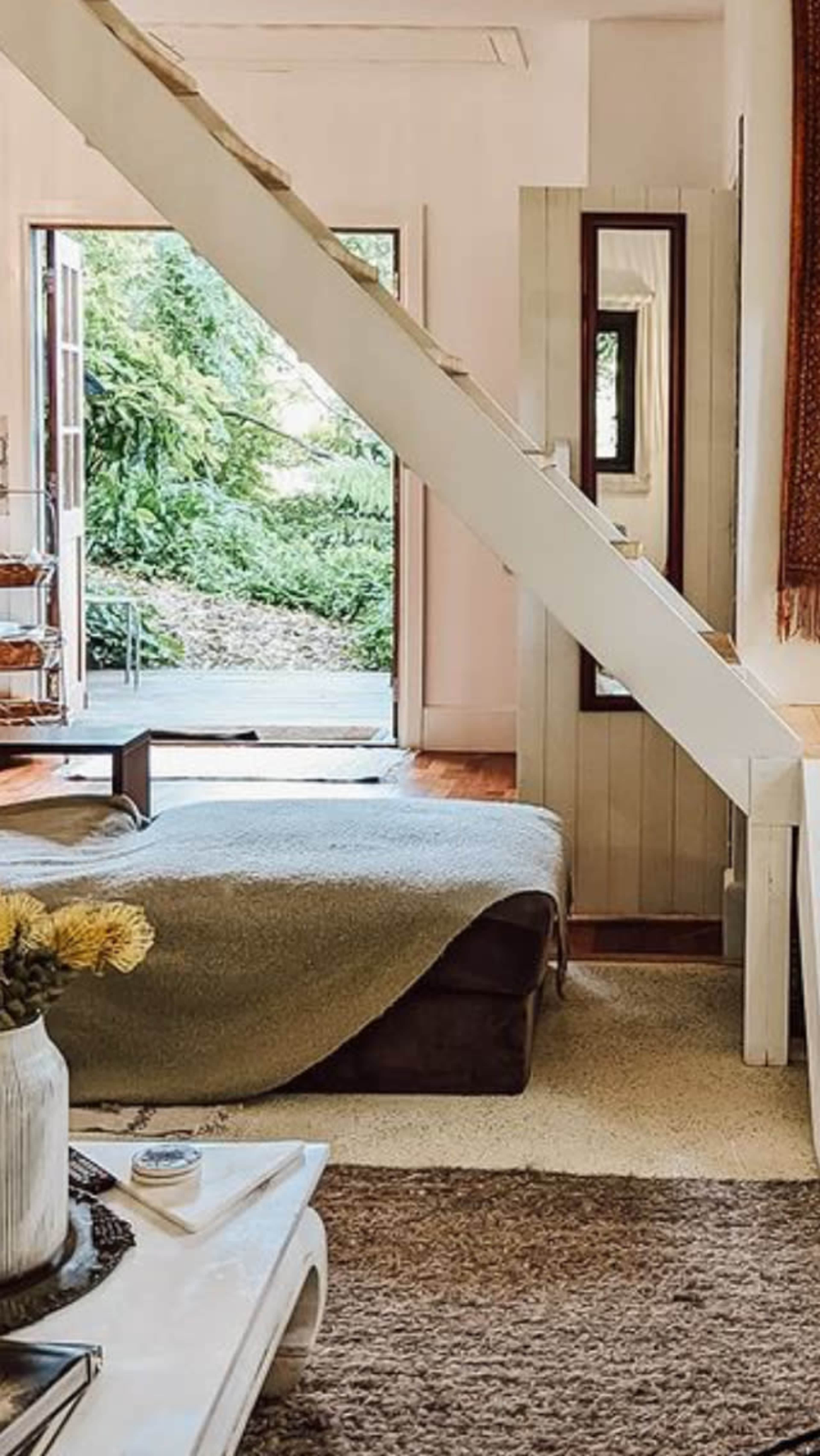 The image shows a cozy living room with a staircase leading to an upper level, a sofa covered with a blanket, and large open doors revealing greenery outside.