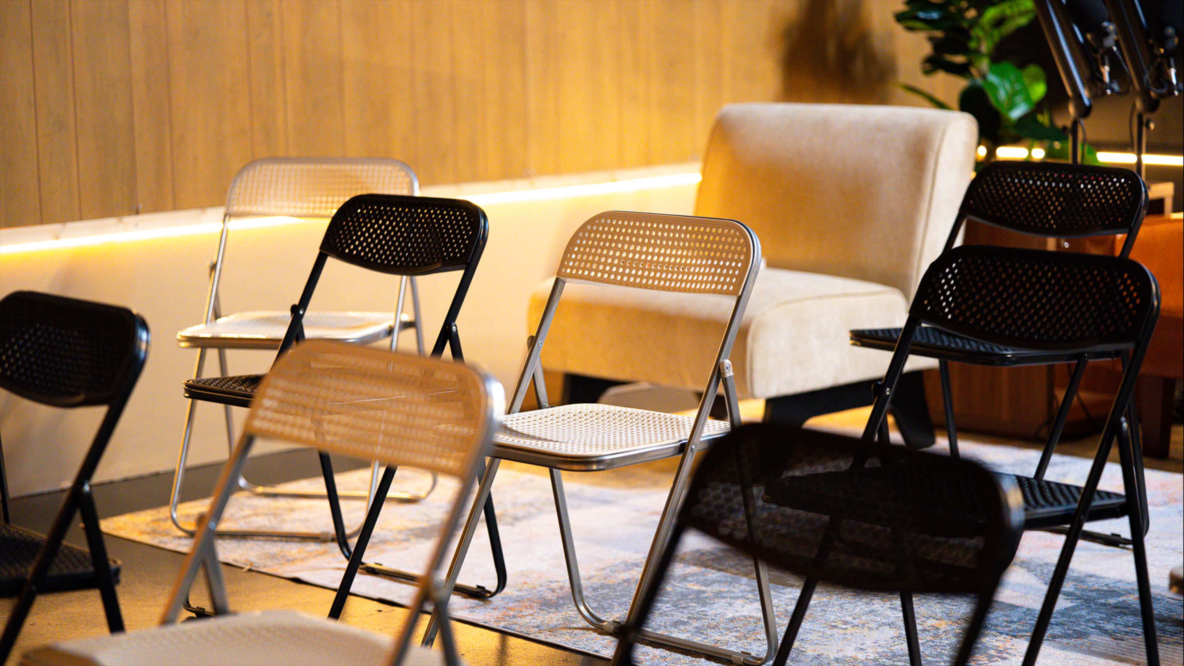 The scene features several folding chairs arranged in a casual setting, with a single upholstered chair and decorative plants in the background.