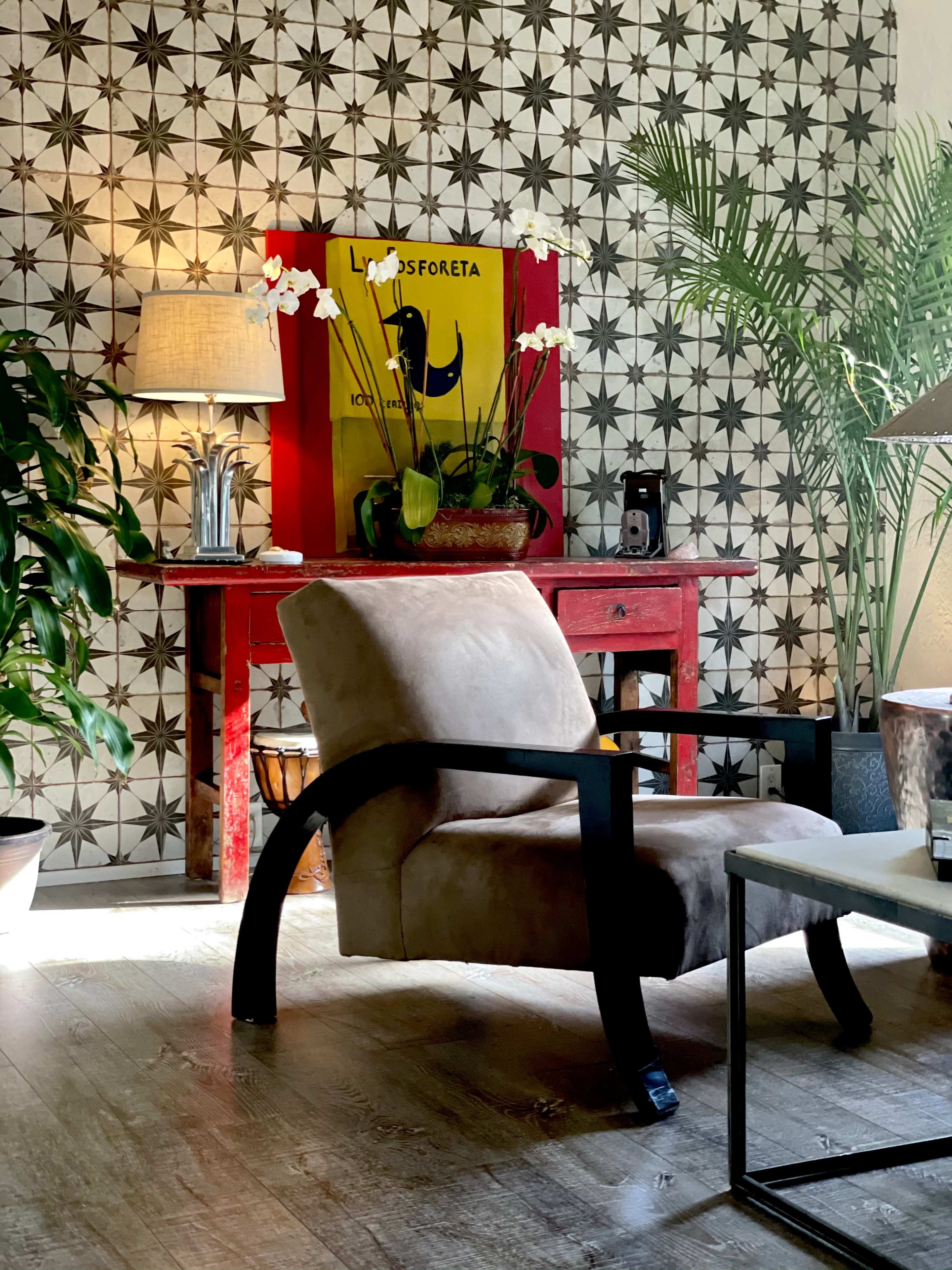 The image shows a cozy living space featuring a beige chair, a red console table adorned with plants, a lamp, and decorative wallpaper with a geometric pattern.