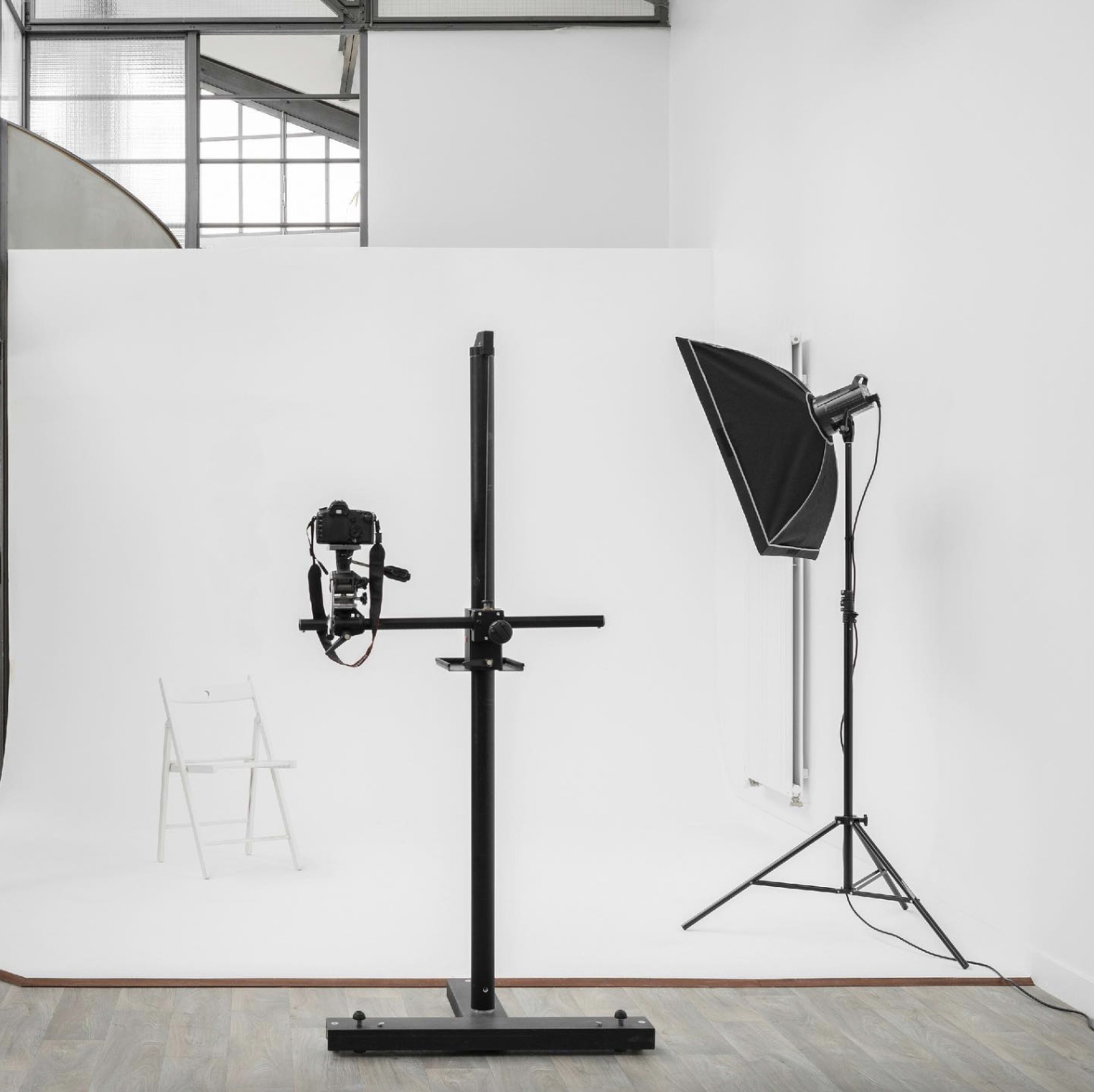 A photography studio with a camera mounted on a vertical stand, a light source on a tripod, and a white backdrop.