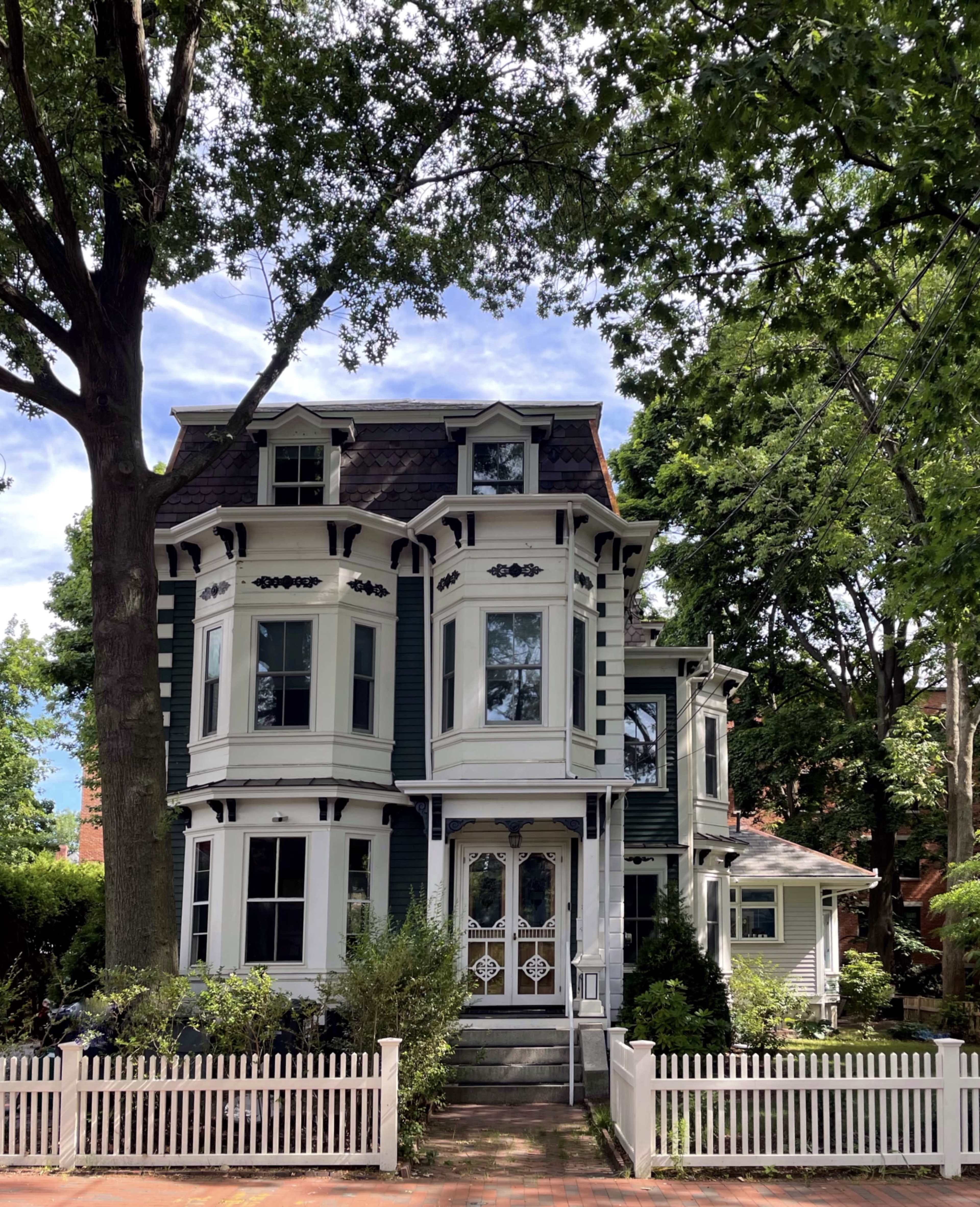 A three-story Victorian-style house with a dark exterior and decorative trim is surrounded by trees and a white picket fence.