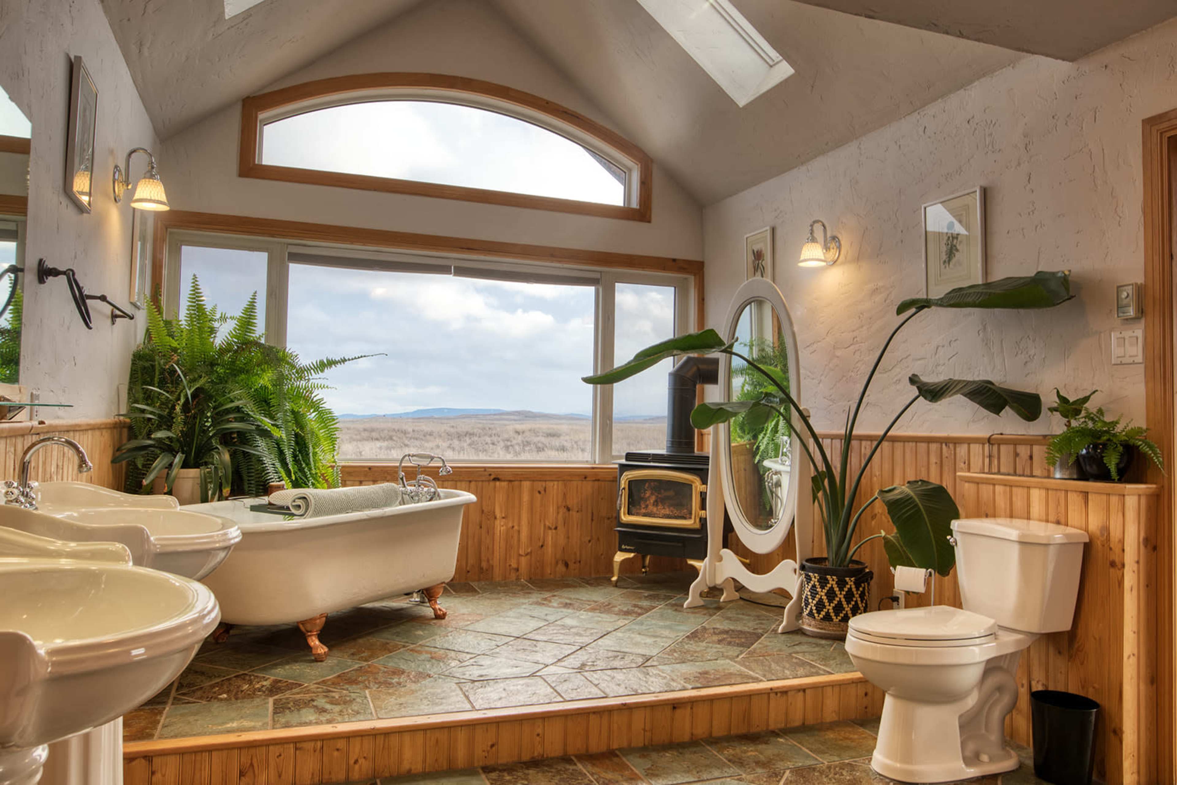 The bathroom features a freestanding bathtub, a large window with a view, plants, and a fireplace against a backdrop of stone flooring and wood accents.