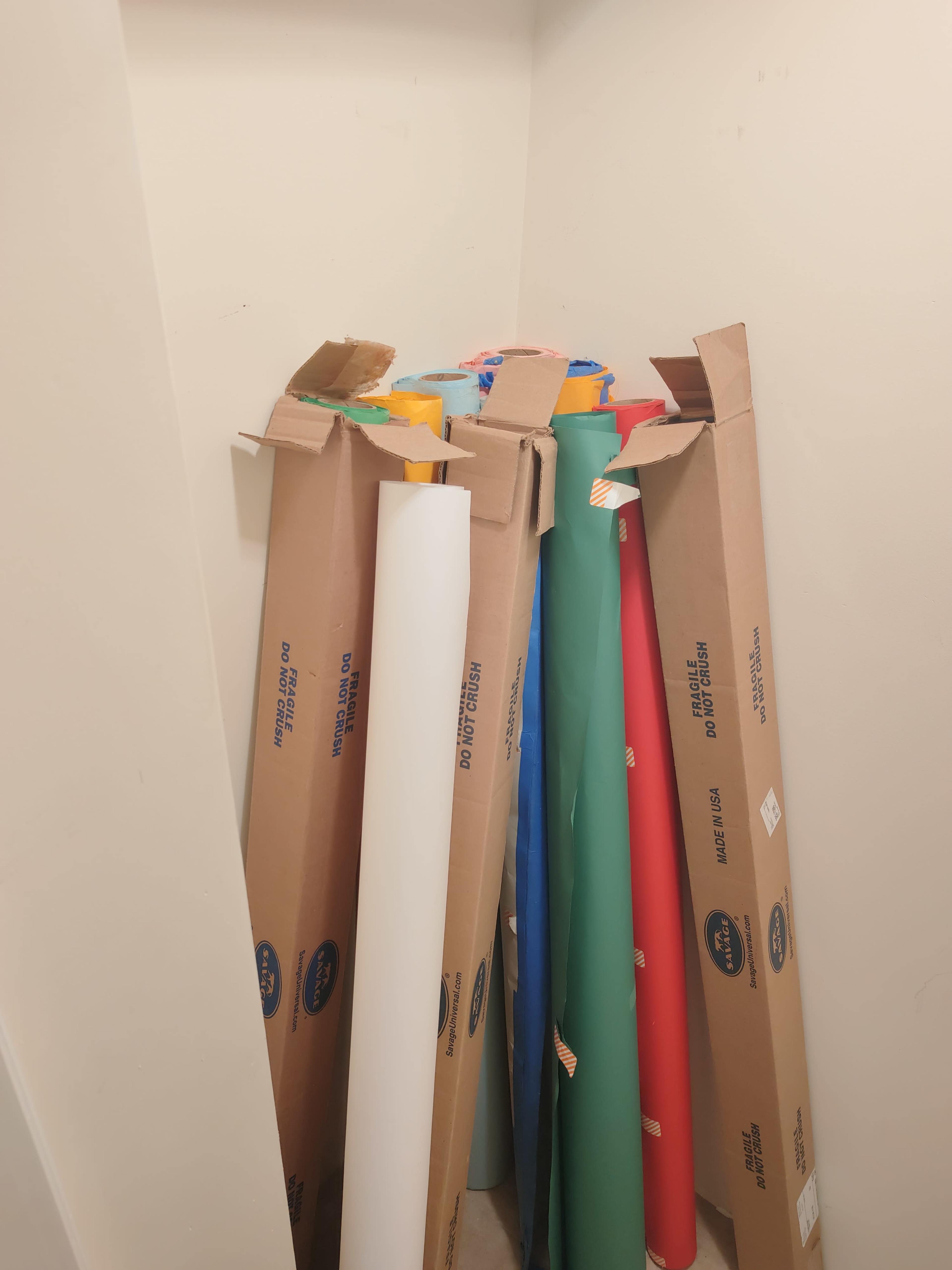 A corner of a room contains several large rolls of colored paper stacked in their cardboard boxes.