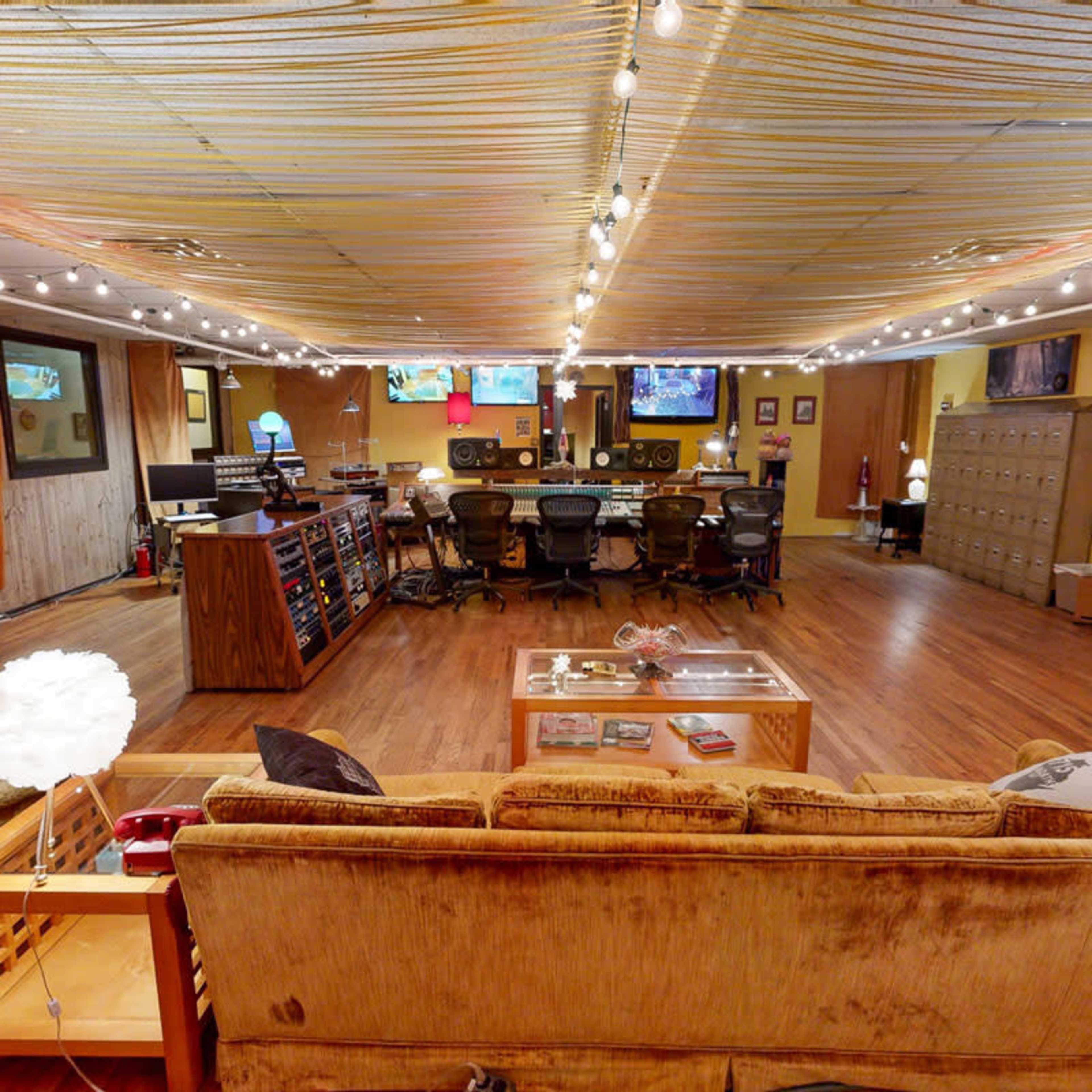 The image shows a spacious, well-lit recording studio with a cozy seating area and various audio equipment arranged along one side.