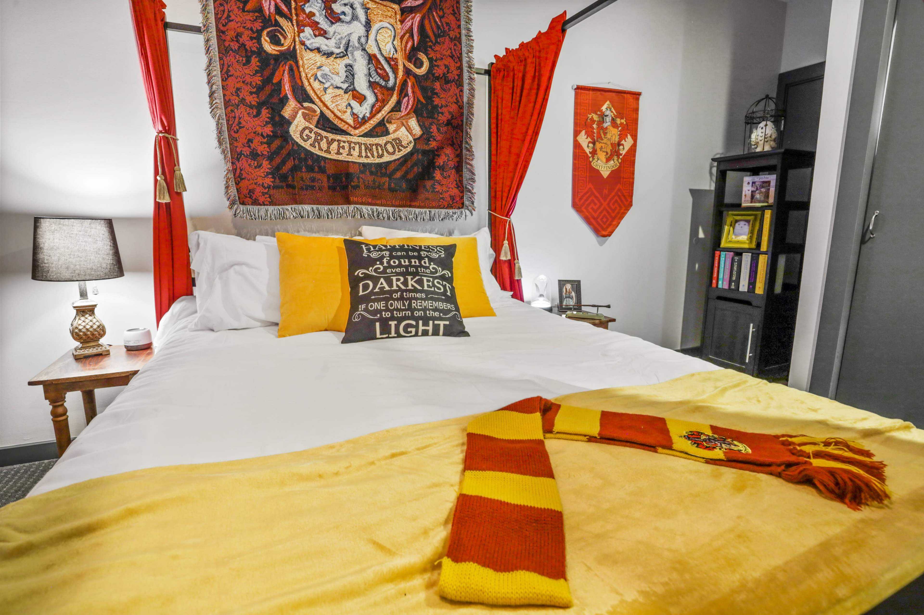 A bedroom features a Gryffindor-themed decor with a large tapestry, colorful bedding, and a lamp on a wooden bedside table.